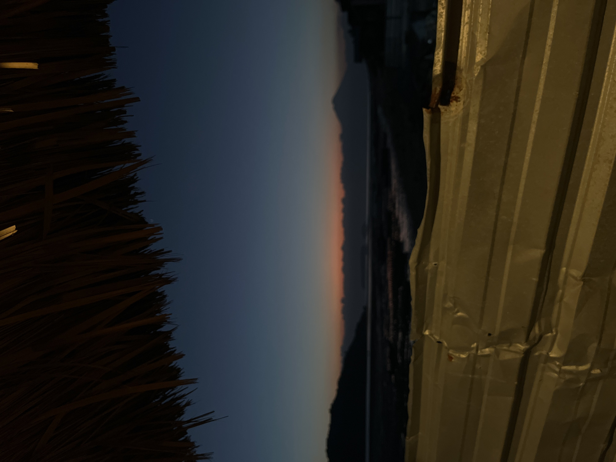 The image depicts a twilight scene overlooking a body of water with a silhouetted mountainous landscape in the distance. The foreground is dominated by a thatched roof and a corrugated metal structure, which appears to be some form of barrier or makeshift enclosure. The sky is a deep gradient of blue, transitioning to a faint band of orange and pink near the horizon, indicating the sun has recently set. The water's surface is dark and slightly reflective, with some indistinct shapes suggesting shallow areas or exposed seabed. The overall mood is tranquil and somewhat rustic, evoking a sense of being in a natural setting with man-made elements providing a partial enclosure. The location is identified as Kertasari, Indonesia, suggesting this is a coastal or lakeside view in that region.