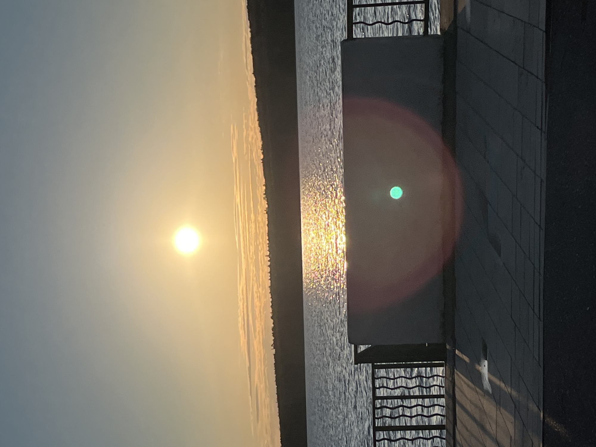 A wide shot, captured in a vertical orientation, depicts a serene sunset or sunrise scene over a body of water, likely a river or lake, in Chaikovsky, Russia. The sky, illuminated by the low-lying sun, displays a gradient of warm golden-orange hues on the left, transitioning to a cooler, hazy blue-gray towards the upper right.
The sun is a brilliant, glowing orb positioned on the left side of the frame, casting a strong, shimmering path of light across the rippling surface of the water. In the distance, a dark, undulating treeline or forested bank marks the opposite shore.
On the right side of the image, a man-made structure, possibly a promenade or pier, extends into the foreground. It features a solid, dark-colored wall or block and a distinctive metal railing with a decorative, wavy pattern. The ground immediately adjacent to this structure appears to be tiled pavement. A prominent circular red and orange lens flare, with a small teal green dot at its center, is visible on the right side, partially obscuring the structure and water. No discernible text is visible in the image.