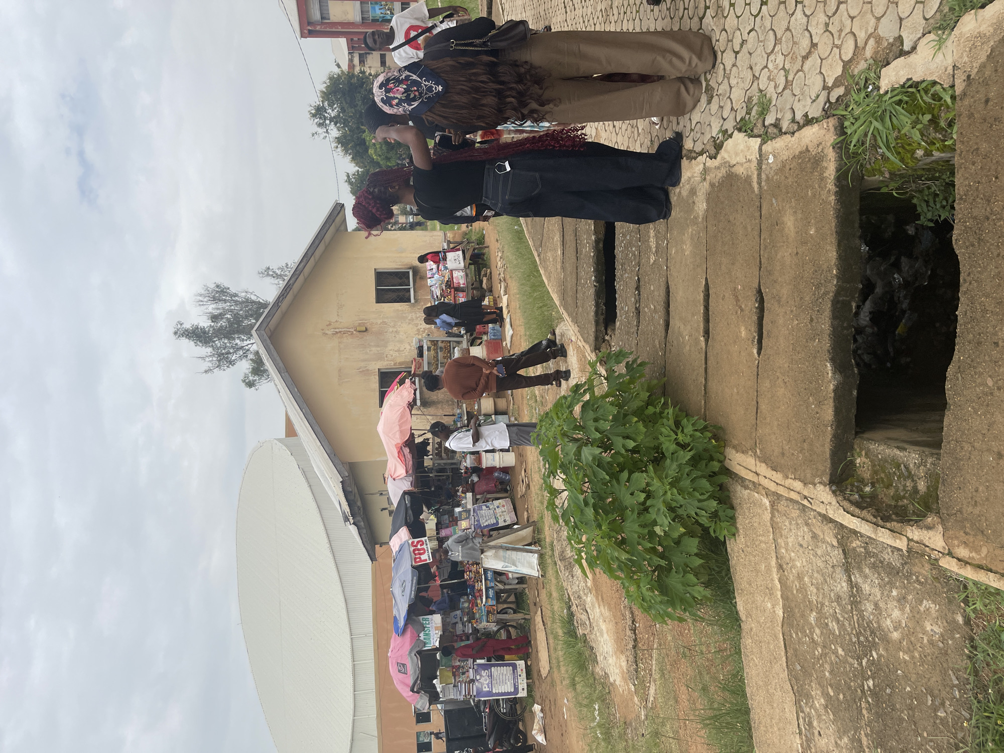 The image depicts an outdoor market scene under a cloudy sky. In the foreground, a concrete drainage channel runs horizontally across the frame, with a large green plant growing to its left. Beyond the channel, a paved area with a circular pattern leads to a market area where several stalls are set up under colorful umbrellas. People are gathered around the stalls, browsing goods, and interacting.
In the background, a light-colored building with a sloping roof is visible. To the left of this building, a large, curved, white structure with a metal roof dominates the upper portion of the image, likely an arena or large hall. The environment appears to be somewhat unkempt, with dirt and patchy grass surrounding the paved areas.
The people visible are dressed in casual clothing. Some individuals are attending to the market stalls, while others appear to be customers. The market seems to be selling various items, as suggested by the goods displayed on tables and shelves. A motorcycle is parked near one of the stalls.
The scene is consistent with an open-air market typically found in urban areas of Nigeria, such as Jos. The combination of market stalls, open spaces, and the presence of both modern and older architectural structures aligns with the urban landscape of the city.
