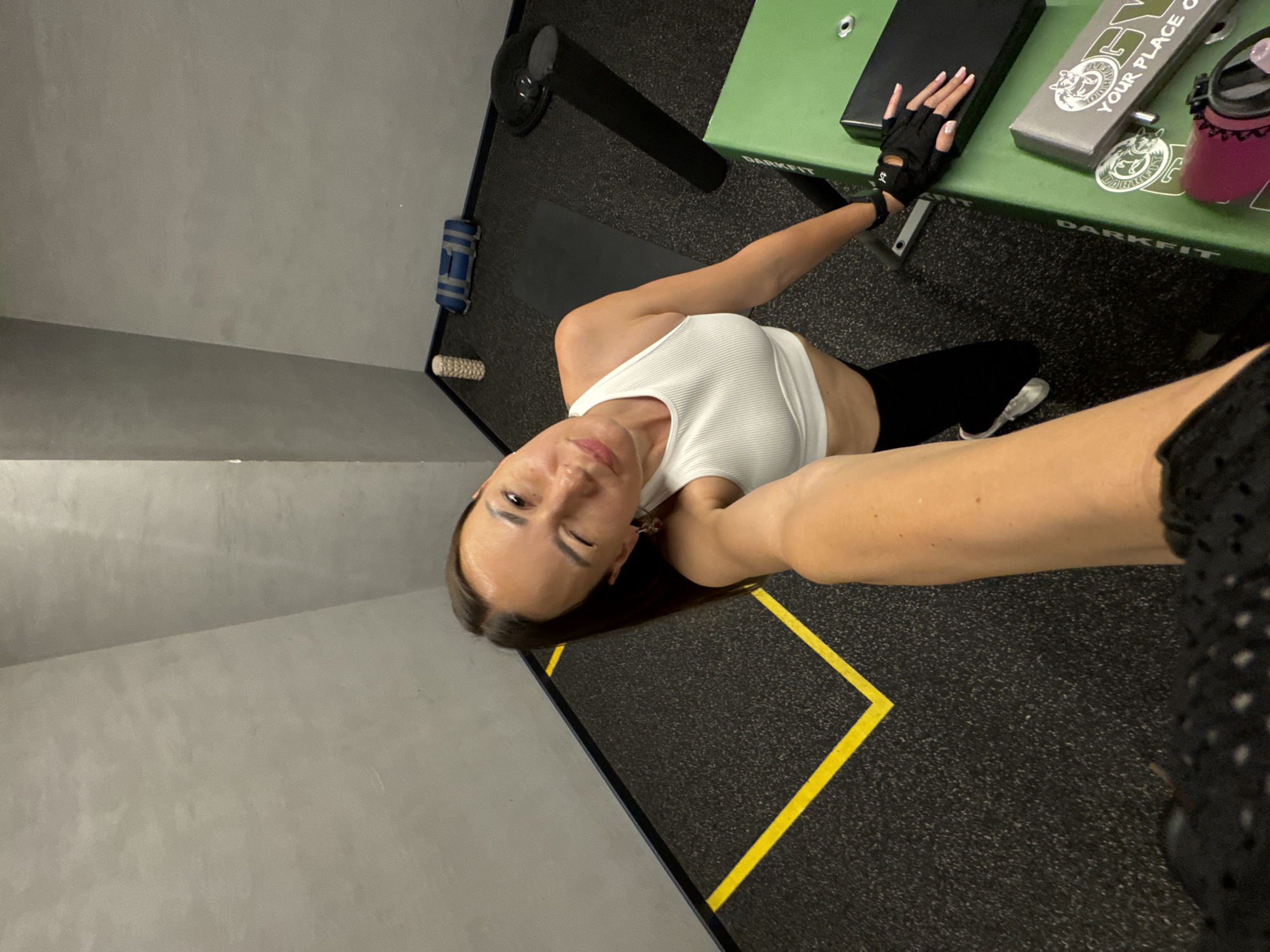 The image shows a woman taking a selfie in a gym. She is wearing a white ribbed tank top and black leggings, with black fingerless gloves on her hands. She has dark hair and is winking at the camera. She is leaning on a green weight bench, which has "DARKFIT" printed on it. Next to her on the bench is a black book or tablet and a grey box with a logo and text that reads "YOUR PLACE O". A pink water bottle is also on the bench. The gym floor is black rubber matting with yellow markings. The walls are a neutral gray color. The scene appears to be indoors, with artificial lighting. The overall mood is energetic and confident, suggesting the woman is about to work out or has just finished.