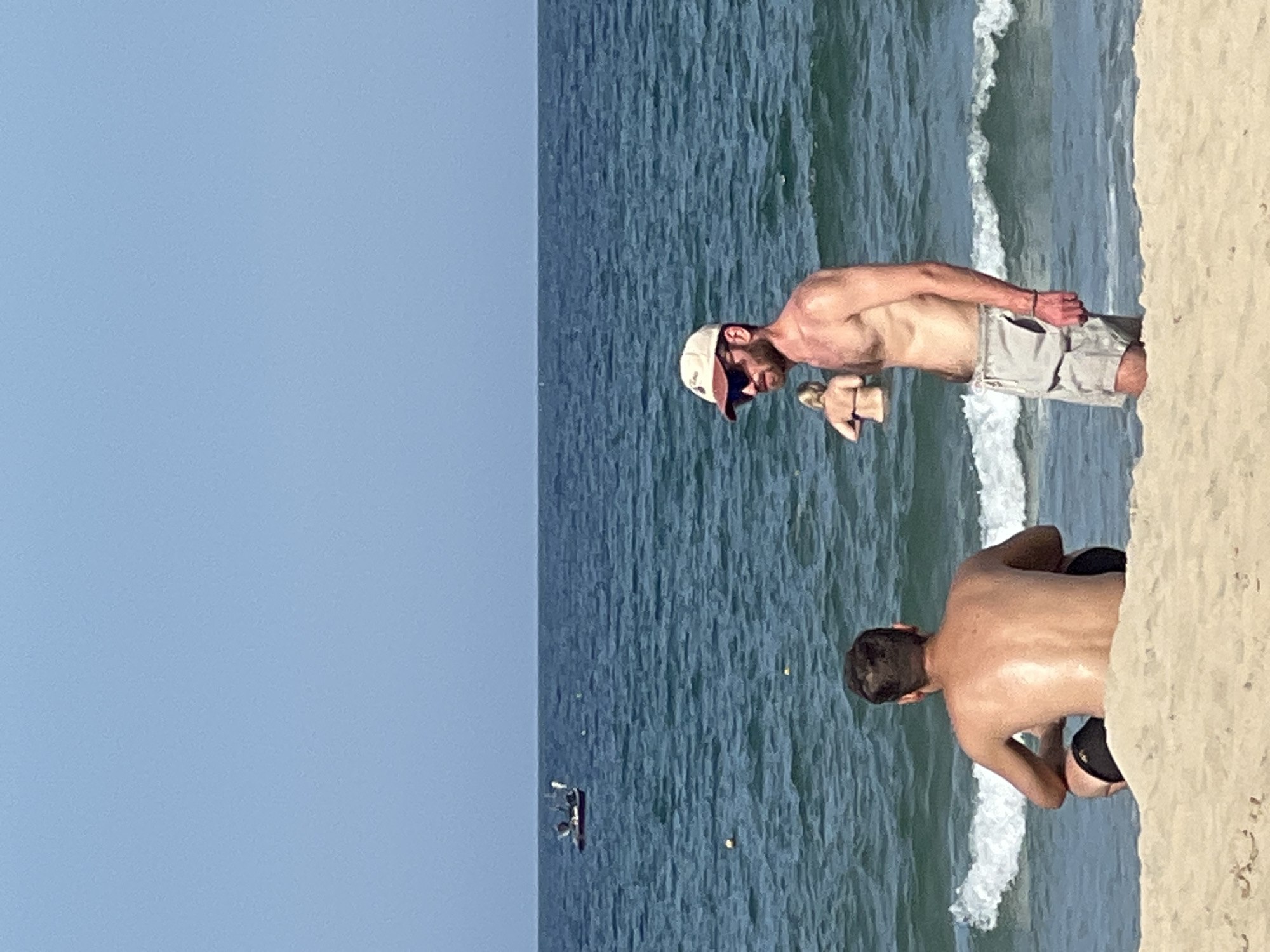 Two shirtless men are on a sandy beach with the ocean in the background. The man on the left is sitting on the sand, facing away from the camera and towards the ocean. He has dark hair and is wearing black swim trunks. The man on the right is standing and facing towards the left. He is wearing a tan baseball cap, sunglasses, and light-colored swim trunks. Behind him, a woman in a bikini is standing in the water. In the distance, a small boat is visible on the ocean. The sky is clear and blue. The scene is likely on a beach in Đà Nẵng, Vietnam, given the characteristic clear blue skies and ocean.