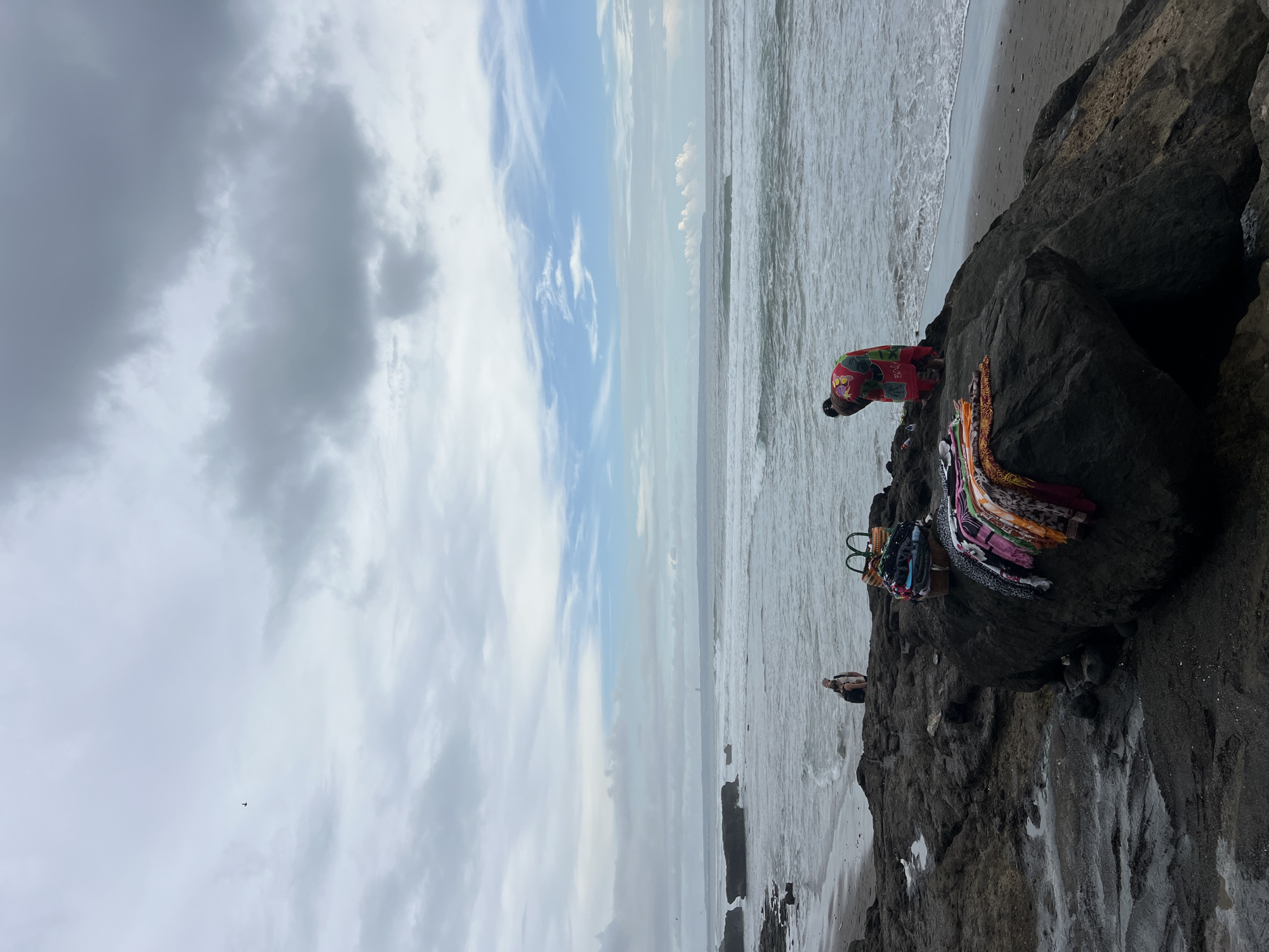 A scenic view of a rocky beach in North Kuta, Indonesia, is captured under a partially cloudy sky. The horizon line separates the expansive sky on the left from the ocean and beach on the right. Large, dark rocks dominate the foreground and mid-ground, extending towards the water.
In the mid-ground, on top of one of these large rocks, a person wearing a vibrant red garment with a colorful pattern (likely floral or traditional) is bending over. Beside them, a collection of colorful textiles, possibly sarongs or scarves, are neatly arranged or stacked. These textiles display a variety of patterns and hues, including pink, green, yellow, orange, and blue. Some items appear to be in simple bags or baskets next to the main stack.
Further to the right and slightly lower on the rocks, another individual is seated, looking out towards the ocean or resting. The sea is a mix of grey and white as waves gently break against the dark sandy shore. The sky above is a blend of light blue and various shades of grey clouds, indicating a daytime setting with diffused light. A small, dark object, possibly an airplane or a bird, is visible in the upper left portion of the sky. No visible text is present in the image. The scene conveys a calm, somewhat overcast day at the beach, possibly depicting a local vendor.