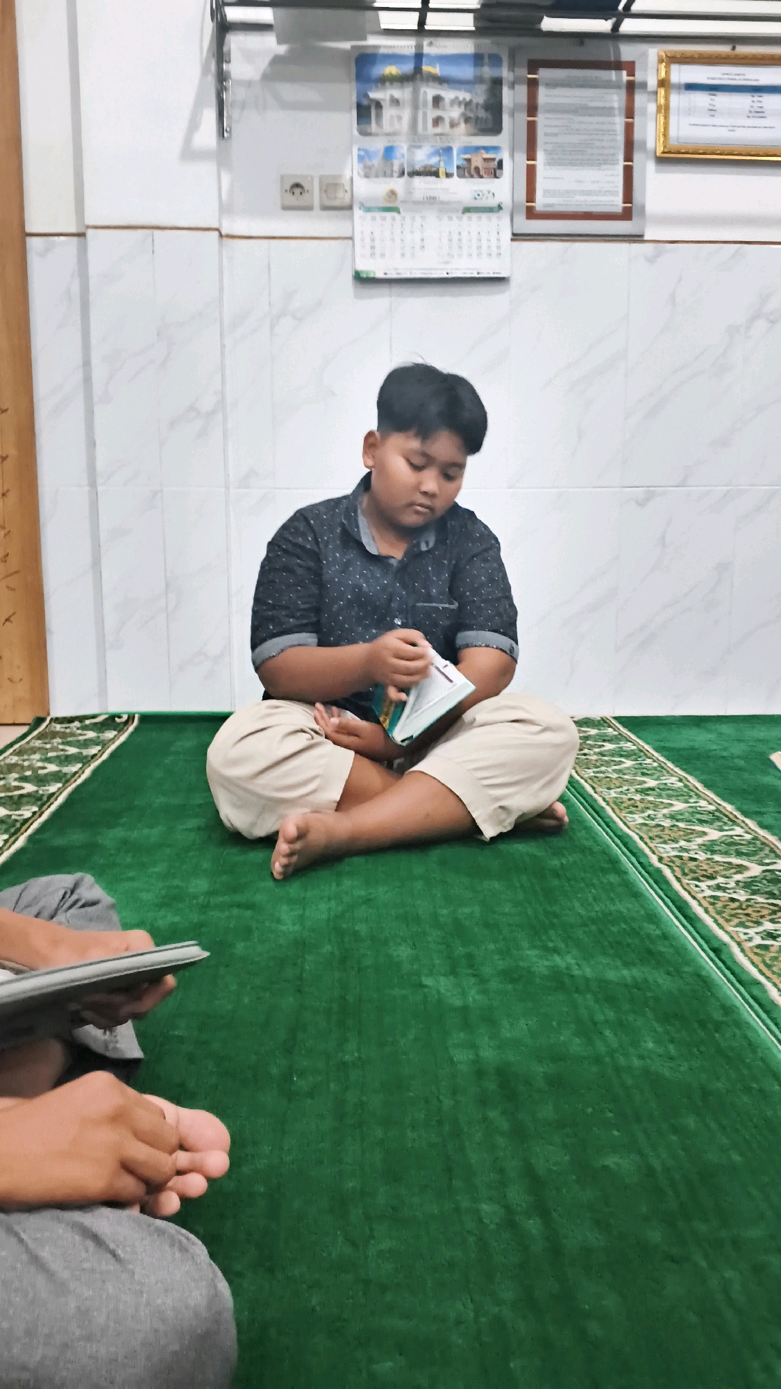 A young boy is sitting cross-legged on a green carpet in what appears to be a mosque or prayer room in Pundungsari, Indonesia. He is wearing a dark patterned short-sleeved shirt and light-colored pants. The boy is looking down and appears to be reading a small, light-colored book. In the foreground, another person's legs and hands are visible, also sitting on the carpet and holding a book. The walls of the room are tiled with a white marble pattern. A calendar with images of mosques is hanging on the wall, along with a framed document. A shelf with some items is mounted above the calendar. The lighting in the room is bright, suggesting it is daytime.