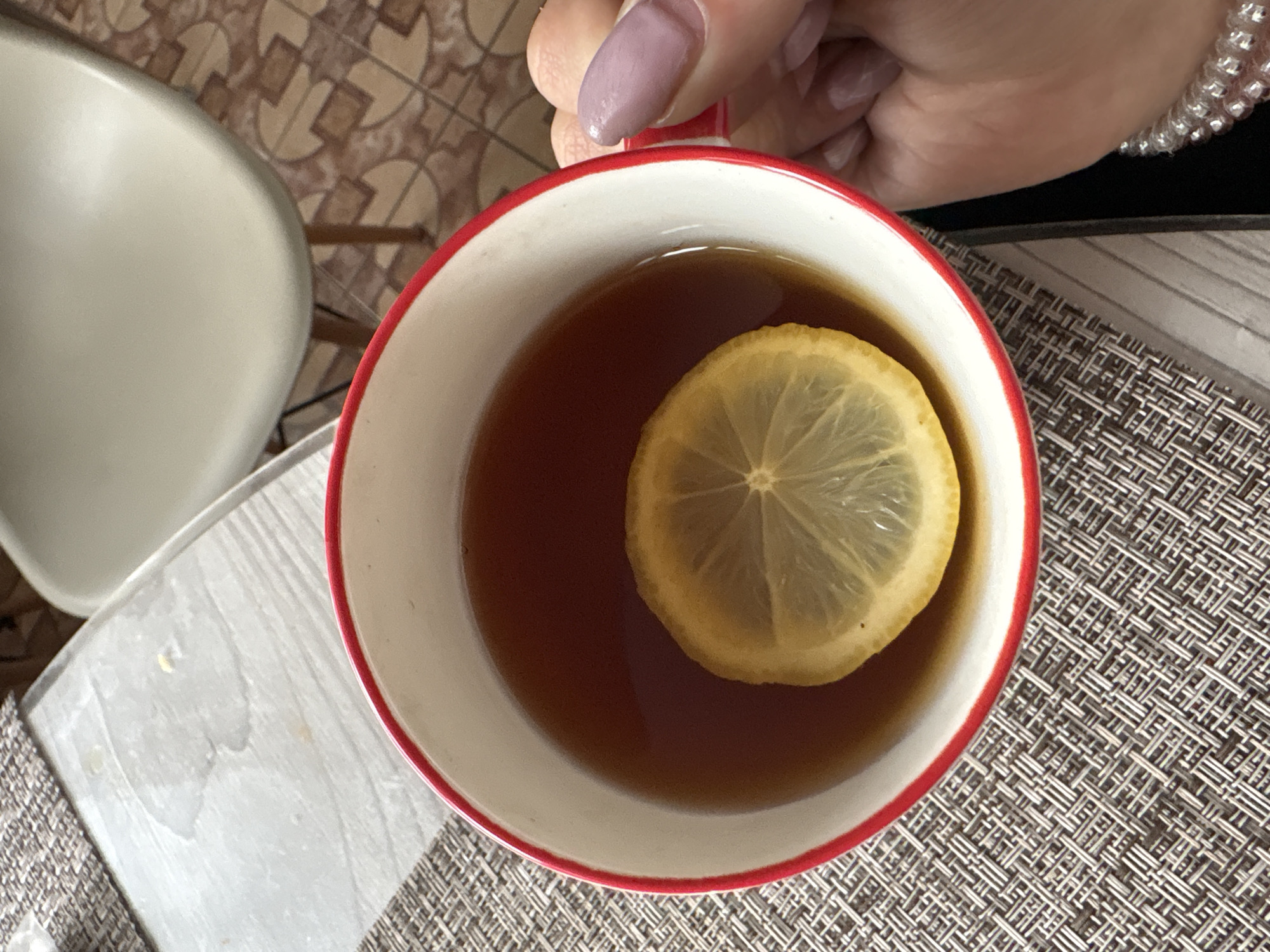 A top-down shot shows a hand holding a red-rimmed white mug filled with tea and a slice of lemon. The mug sits on a woven placemat. Part of a chair is visible on the left. The photo location is unknown and cannot be determined as Прикамский is a rural settlement without sufficient geographic markers visible in the image.