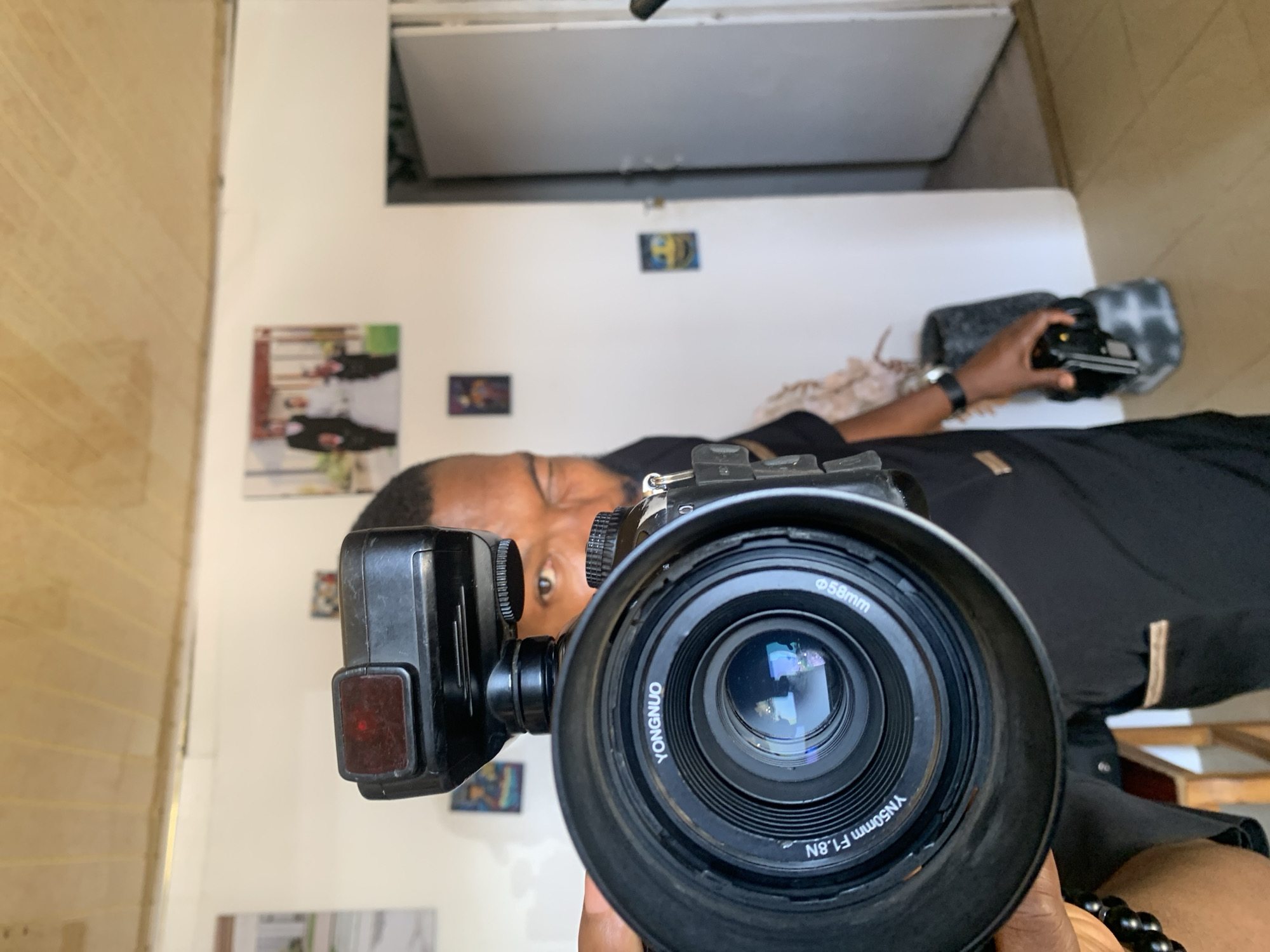 The image, rotated 90 degrees counter-clockwise, captures a first-person perspective of a photographer holding a camera. The primary focus is a black DSLR or mirrorless camera with a large black lens, featuring a lens hood. Visible text on the lens indicates it is a "YONGNUO YN50mm F1.8N" with a "ø58mm" filter thread. An external flash unit, with a red sensor window, is mounted on the camera's hot shoe to the left of the lens.
Behind the camera, the photographer's face is partially visible, showing dark eyes and forehead as they look directly into the lens. The person has dark skin and short dark hair/beard, wearing a black shirt. Their right hand holds the camera, while their left hand is seen in the background holding a smaller black electronic device, possibly a remote trigger, resting on a grey, textured object. A black watch adorns their left wrist, and a black beaded bracelet is on their right wrist.
The setting is an indoor room in Jos, Nigeria. The background consists of a plain white wall displaying several small framed pictures or prints, one of which is brightly colored. To the far left, a segment of a wall with light wood-paneled or tiled surfaces is visible. Above, a white ceiling or the underside of an overhead fixture is present. The scene suggests a self-portrait or a setup for taking a photograph, with the photographer directly engaging with their equipment.