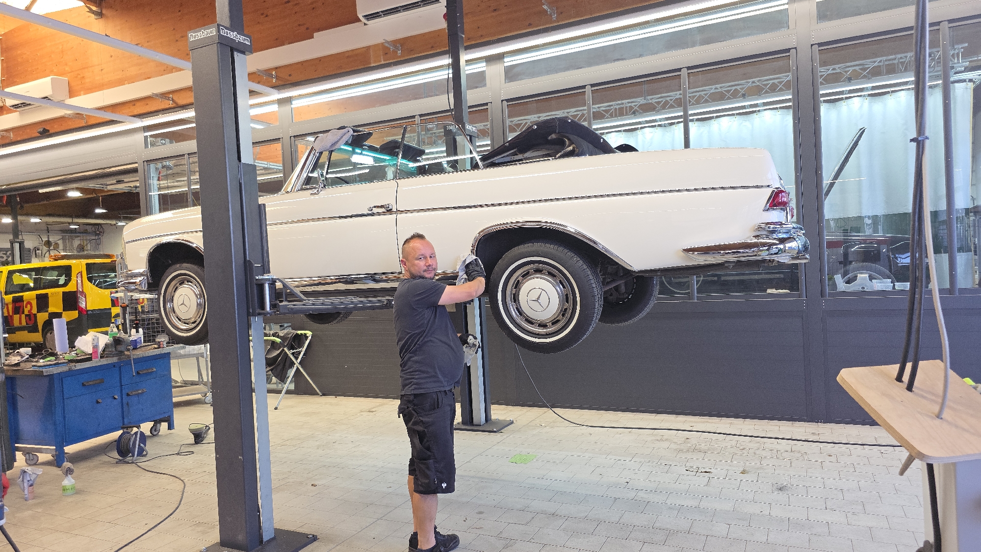 In a car repair shop, a man is working on a white convertible Mercedes-Benz. The car is raised on a hydraulic lift. The man is wearing a black t-shirt, black shorts, and black gloves. He is cleaning the wheel arch of the car. There are tools and equipment around the shop, including a yellow and black service van in the background. The shop has wooden paneled walls and ceiling, and fluorescent lighting. The floor is tiled. A wooden table with a metal frame is in the foreground.