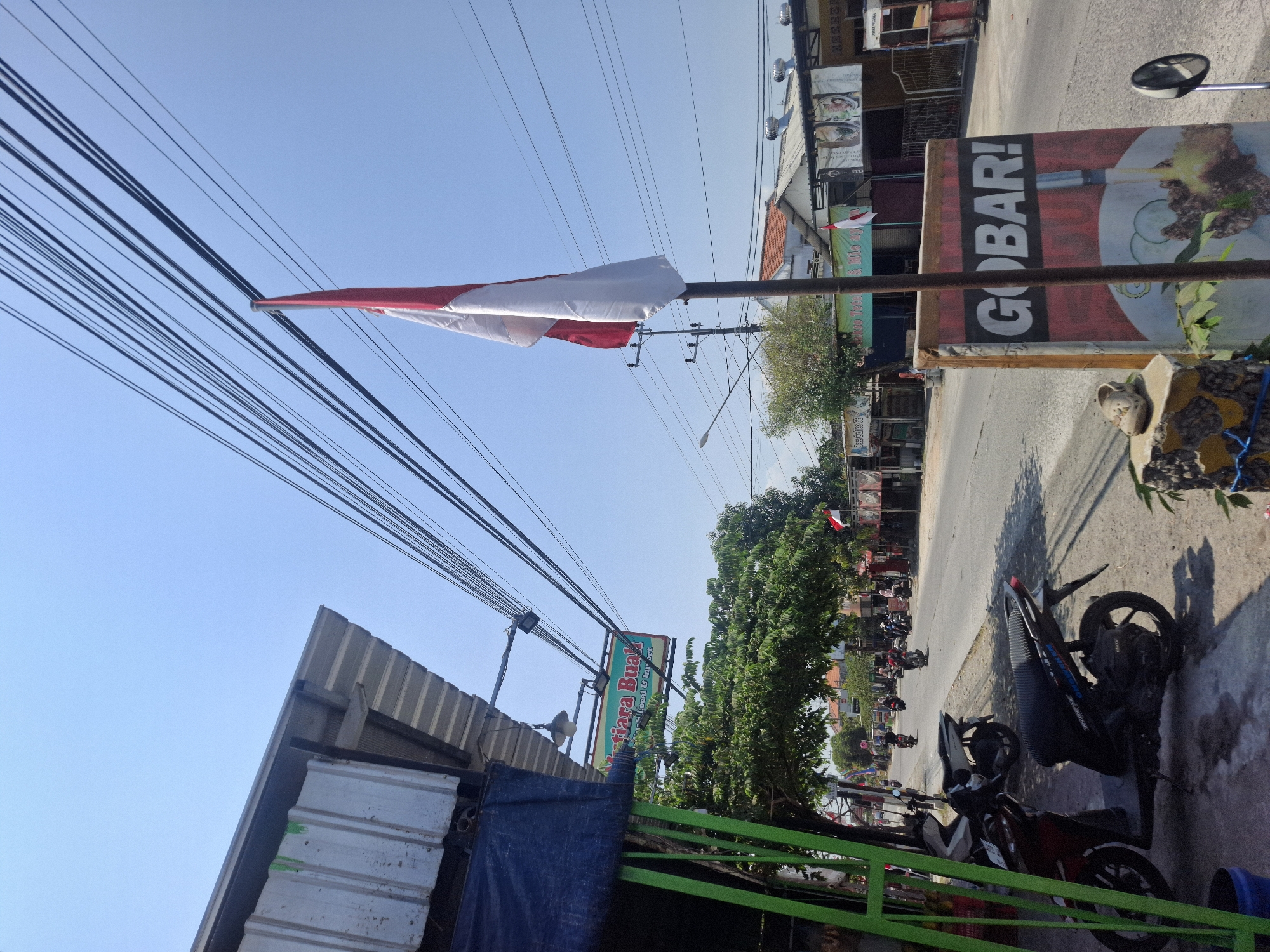 The image depicts a street scene in Sumberwudi, Indonesia, on a clear, sunny day. The foreground is dominated by a tall flagpole displaying the Indonesian flag, which is red and white. To the left of the flag is a sign with the text "GOBAR!" and an image of food. Further to the left, a building with a corrugated metal roof and a blue tarp overhang is visible, along with a green metal structure. Several motorcycles are parked along the side of the road.
The road itself stretches into the distance, lined with various buildings and shops. There are several trees along the roadside, providing some shade. Multiple Indonesian flags are also visible flying from poles in front of some of the establishments. A few people can be seen on motorcycles in the middle ground of the image, traveling along the road. Overhead, a complex network of electrical wires crisscrosses the sky. The overall impression is a typical street view of a commercial area in Indonesia.