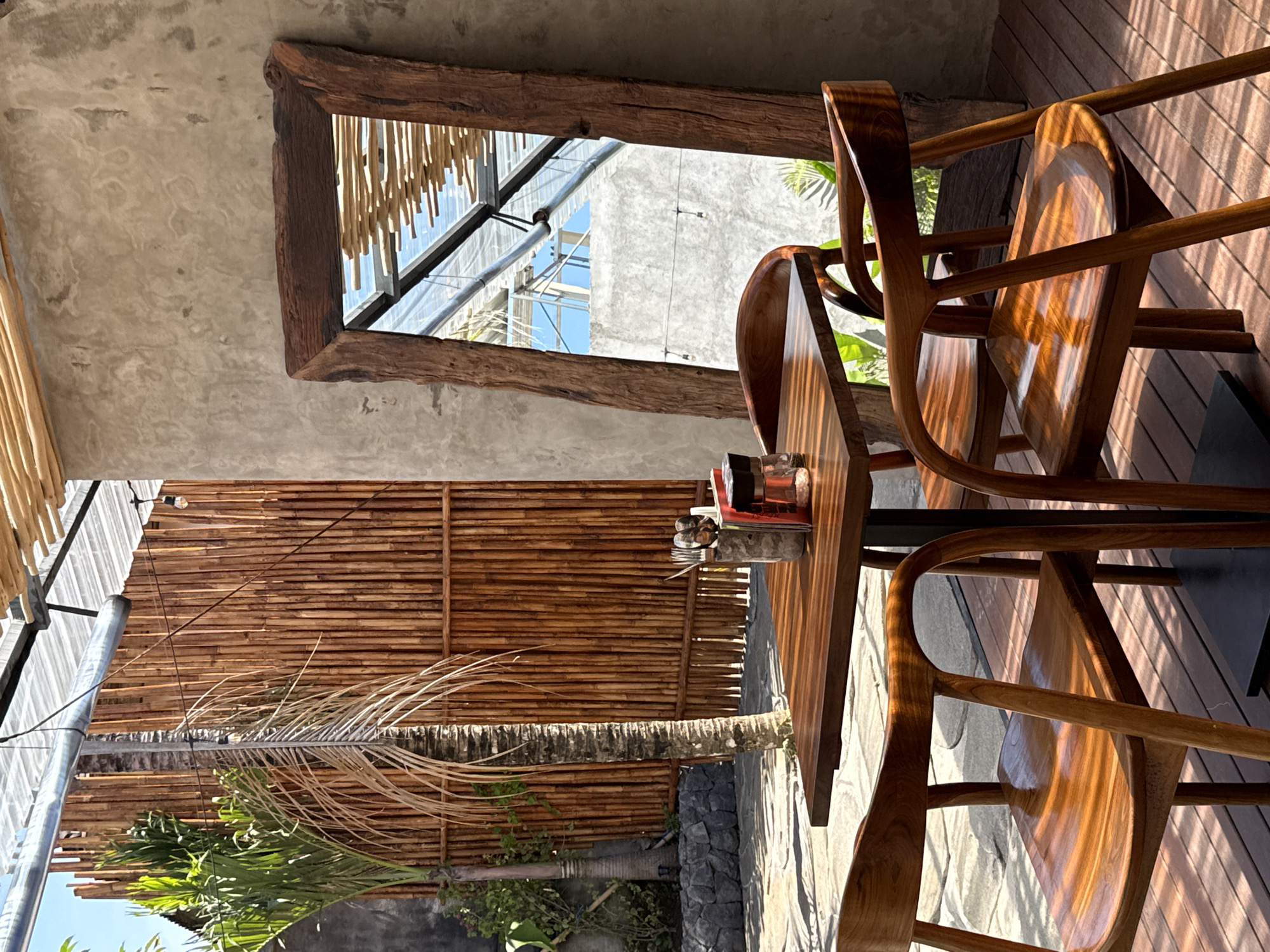 The scene is outdoors, appearing to be in a covered patio or cafe in Kuta Selatan, Indonesia. The setting includes wooden furniture, specifically a wooden table and chairs, positioned on a wooden deck. The area is enclosed by a bamboo fence. A wooden frame provides a partial view of a building across a narrow alley. Natural light filters through the canopy and creates shadows on the deck. A palm tree is visible in front of the bamboo fence. The sky can be seen in a sliver above the tree. Items are on the table.