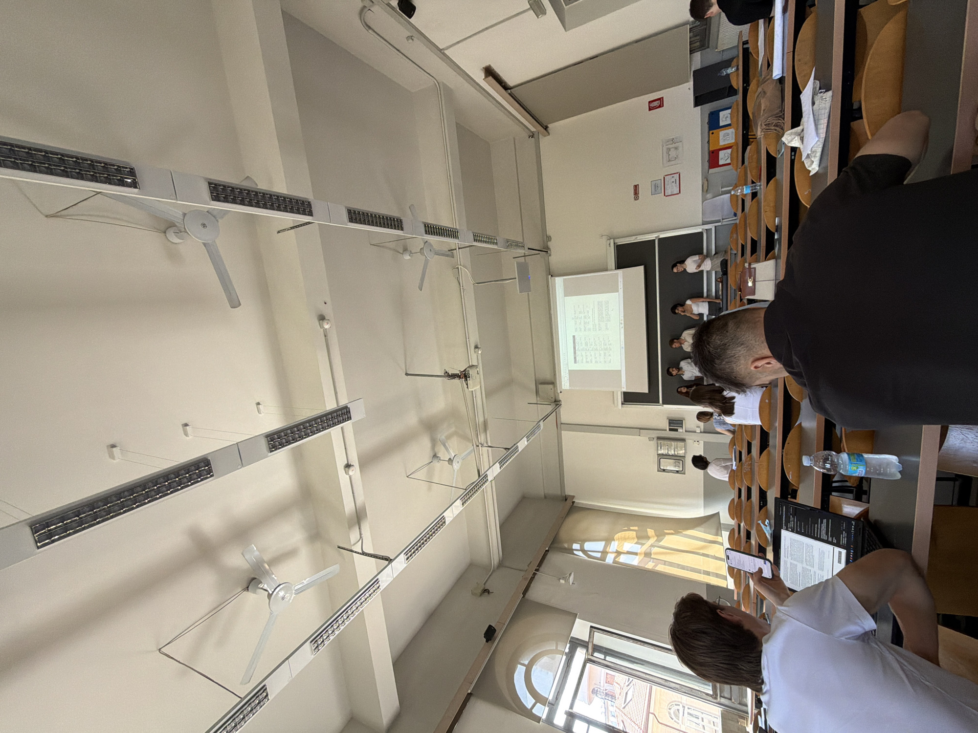 The image depicts an indoor lecture hall at an educational institution, likely a university, in Milan, Italy. The scene is viewed from a student's perspective, looking towards the front of the room.
**Environment:**
The lecture hall features a high, white ceiling with multiple fluorescent light fixtures and three white ceiling fans, some of which are positioned centrally and others towards the sides. The walls are also white. A large projection screen displaying text is mounted on the front wall. Rows of tiered wooden desks with cushioned seats are arranged in the room. Large arched windows with blinds are visible on the left side, allowing natural light to enter and offering a partial view of buildings with red-tiled roofs, characteristic of Milan's architecture. The overall impression is one of a functional, somewhat dated, academic setting.
**People:**
In the foreground, two students are seated at desks. The student on the left, seen from the back and right side, is wearing a white t-shirt and is looking at their phone while using a laptop. The student on the right, also seen from the back, is wearing a black t-shirt and appears to be focused on something on their desk. In the middle ground, several students are seated at desks, some facing forward, others turned to the side. At the front of the room, a group of individuals, possibly instructors and students, are standing near the projection screen. One person is speaking at a podium.
**Objects:**
Prominently displayed are the desks, chairs, ceiling fans, light fixtures, and the projection screen. On the desks, laptops, phones, and a bottle of water are visible. On the front wall, various notices and posters are affixed. A projector is mounted on the ceiling.
**Actions and Interactions:**
Students are engaged in listening to a lecture, using electronic devices, and preparing for or taking notes. The individuals at the front appear to be presenting or teaching. The overall atmosphere suggests a typical academic lecture or presentation.
**Specific Details:**
The presence of ceiling fans suggests the lecture hall may not have air conditioning, or it is not in use. The architecture visible through the windows, with the red-tiled roofs and the style of the buildings, is consistent with urban architecture found in Milan. The signage on the wall, although not clearly legible, appears to be in Italian, further supporting the location.