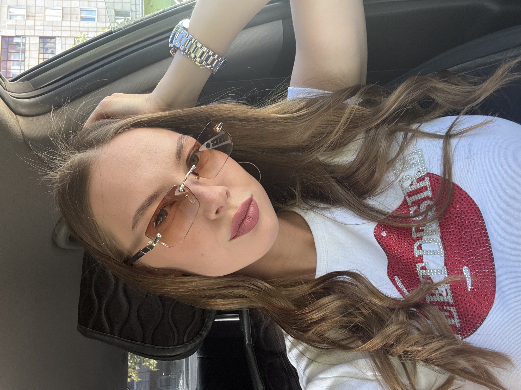 A young woman with long brown wavy hair is in the driver's seat of a car, looking at the camera. She is wearing a white t-shirt with a large red sequined lips graphic and the text "LET'S DESIRE" below it, rimless sunglasses with amber lenses, and a silver wristwatch on her left wrist. Her right arm is raised, with her hand resting on the back of her head. The background shows the interior of the car, including a dark gray headrest and the window, through which a residential building with windows can be seen. The scene appears to be in a city, potentially Perm, Russia, given the architectural style of the building.