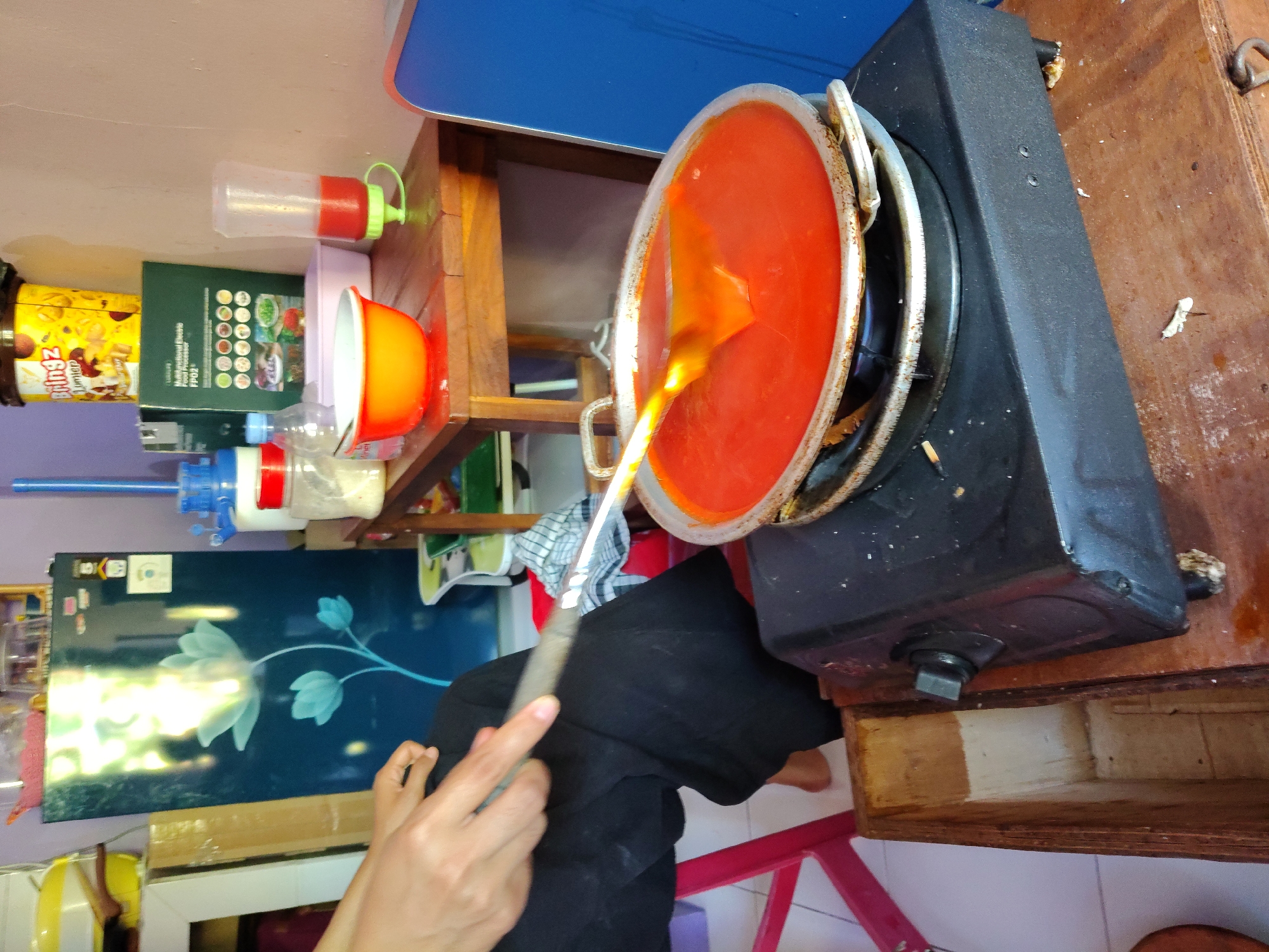 A close-up, eye-level shot captures a kitchen scene. In the foreground, a person's hands, wearing black clothing, are seen on the left. They are holding a spatula and stirring a large, flat, silver pan filled with a vibrant red, semi-liquid substance. The pan is situated on a black electric stove. The stove is placed on a rustic wooden surface, which also has some white debris scattered on it. Behind the stove, a portion of a wooden box is visible.
In the midground, to the right of the stove, a light brown wooden table holds several items: an orange bowl, a plastic squeeze bottle with red liquid, and a clear plastic container with a white lid. Behind the table, a dark blue refrigerator with a floral design is prominent. To the right of the refrigerator, a blue chair or seat is partially visible. The background appears to be a wall painted in a light purple hue. The overall environment suggests a domestic or small-scale food preparation setting. The scene is likely located in a residential kitchen in the city of Surabaya, Indonesia, given the context provided.