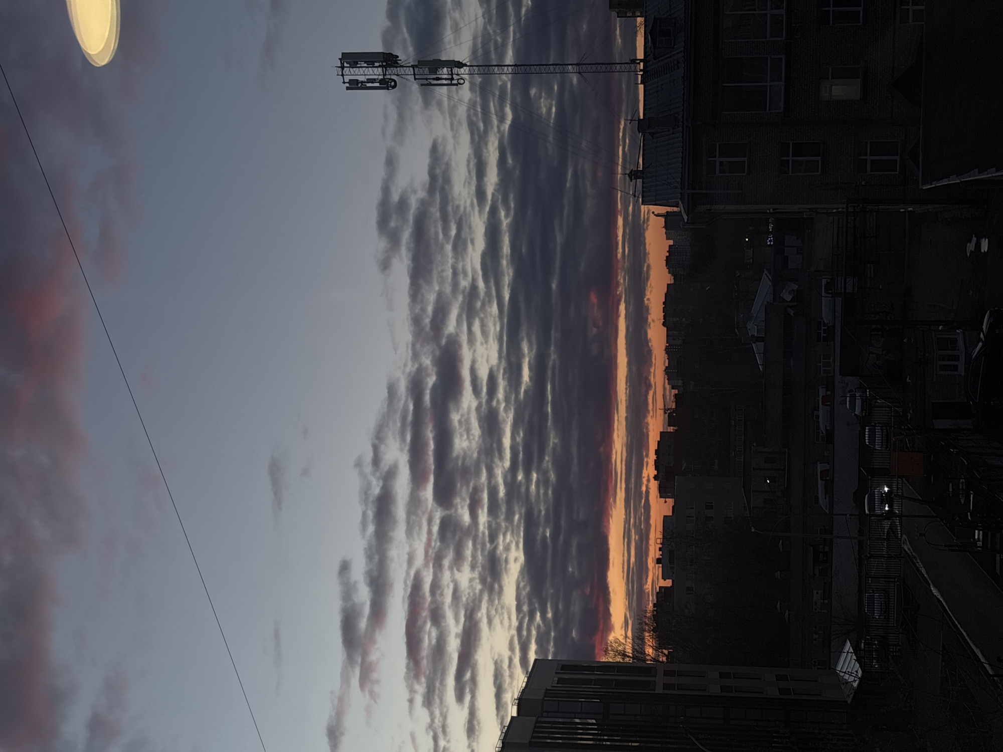 This is an outdoor shot taken in Perm, Russia. The image is oriented vertically. A street lamp appears in the upper-left corner. A large cloud formation stretches vertically across the middle of the image. Beyond this cloud formation is a strip of orange sky. The right side of the image contains several buildings. Cars and street lights are visible. A metal structure sits atop one of the buildings. A power line runs across the left side of the sky.