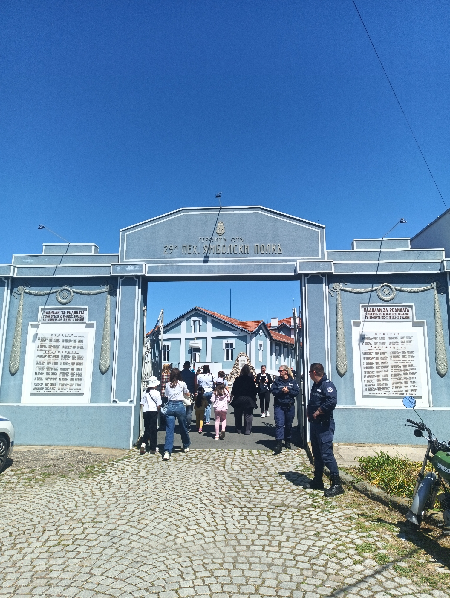 The image captures the entrance gate to the "Heroi ot 29-ti Peh. Yambolski Polk" (Heroes of the 29th Infantry Yambol Regiment) monument in Yambol, Bulgaria. The light blue structure features a central archway with the regiment's name inscribed above. Plaques flank either side of the archway, bearing names of fallen soldiers "Padnali za Rodinata" (Fallen for the Motherland) from the Balkan Wars (1912-13) and World War I (1915-18).
Several people are seen passing through the gate, including a family with a child. A uniformed police officer stands on the right side of the entrance. A Simson motorcycle is parked near the officer. The ground is paved with cobblestones. A light blue building can be seen through the gate. The sky is clear and blue.