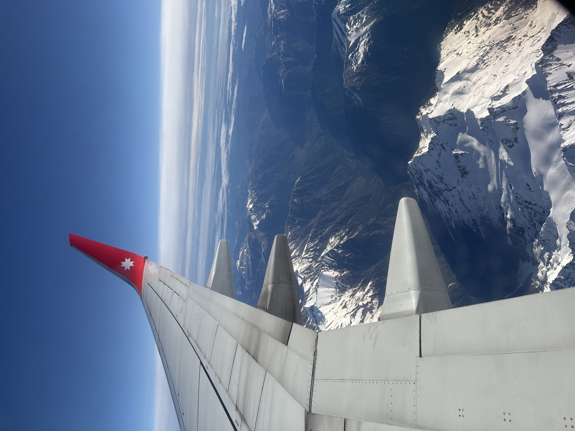 The image shows an airplane wing with a red tip bearing a white star symbol, presumably the Australian flag. The wing is viewed from inside the airplane, with the blue sky and the curvature of the Earth visible to the left of the wing. To the right, mountain ranges are visible, partially covered in snow. There is no evidence to suggest the location is Sochi, Russia.