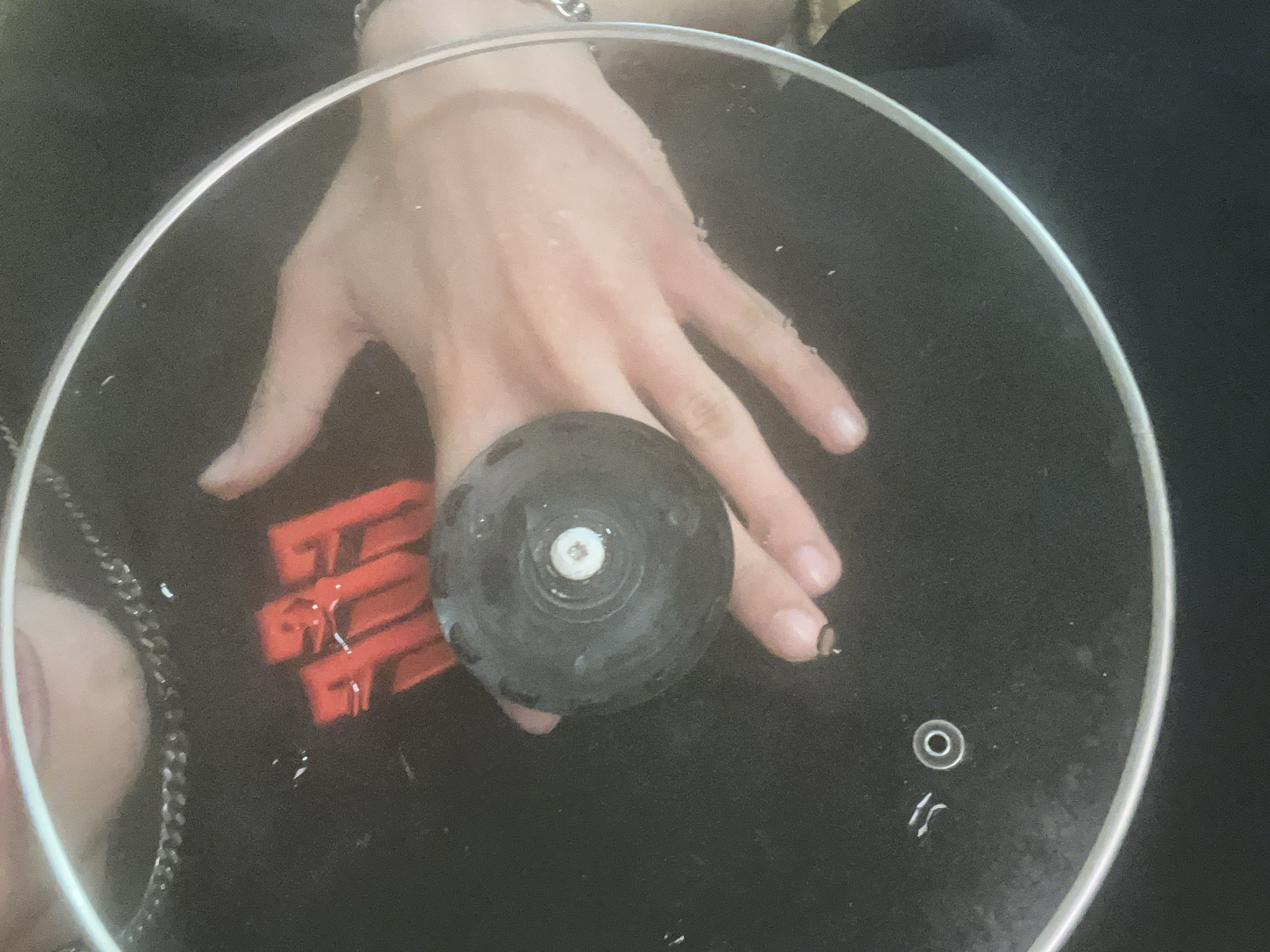 A hand with a bracelet rests on a clear glass lid with a metal rim and black knob. The lid is lying on a black surface. Red letters are partially visible under the hand. A silver chain lies to the left. The setting is indoors. The location is not discernible from the provided image and cannot be determined to be Sofia, Bulgaria.