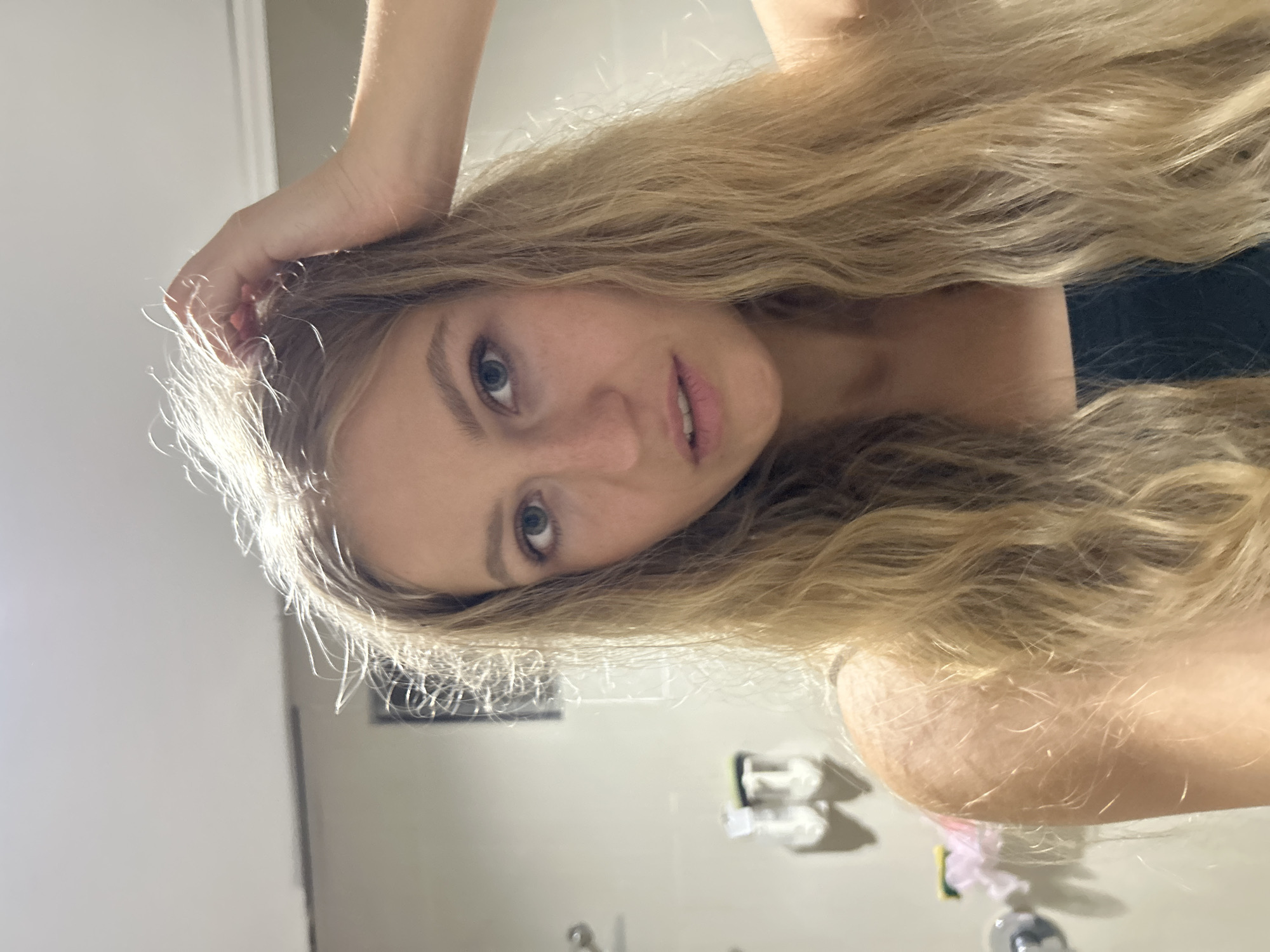 A blonde woman is posing indoors. She is facing the camera and has her hand on her head. The room appears to be a bathroom with a sink and toiletries visible in the background. There is no evidence to indicate a location in Kuta Selatan, Indonesia.