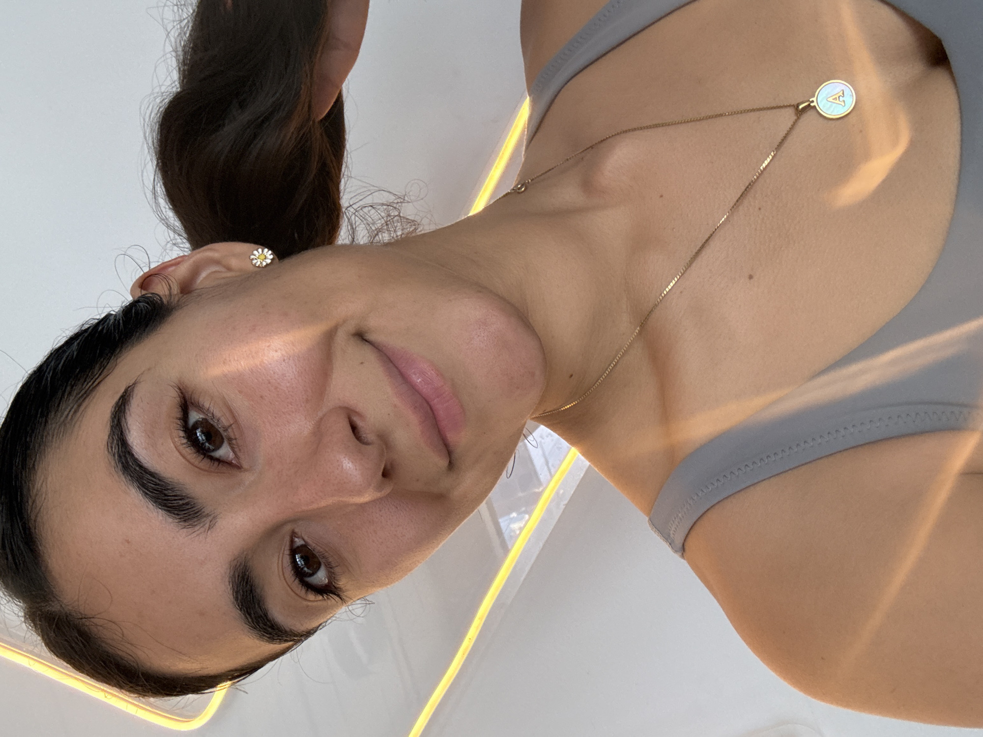 A close-up, rotated portrait captures a young woman from a low angle, revealing her face and upper body against a bright, minimalist background. She has dark hair tied back, dark eyes, and full eyebrows, offering a gentle, content smile with light pink lips as she looks towards the camera.
She is dressed in a light gray top, possibly a sports bra, visible across her shoulder and upper chest. Her accessories include a small white and yellow daisy stud earring in her right ear and a delicate gold chain necklace. The necklace features a circular pendant with a gold rim and an iridescent, possibly mother-of-pearl, center prominently displaying the capital letter "A."
The background is predominantly white or very light, with distinct, linear yellow lights running across the frame, suggesting an indoor setting with modern, illuminated features. The scene takes place in Puebla, Mexico. The lighting is bright, creating a clean and focused image of the subject.