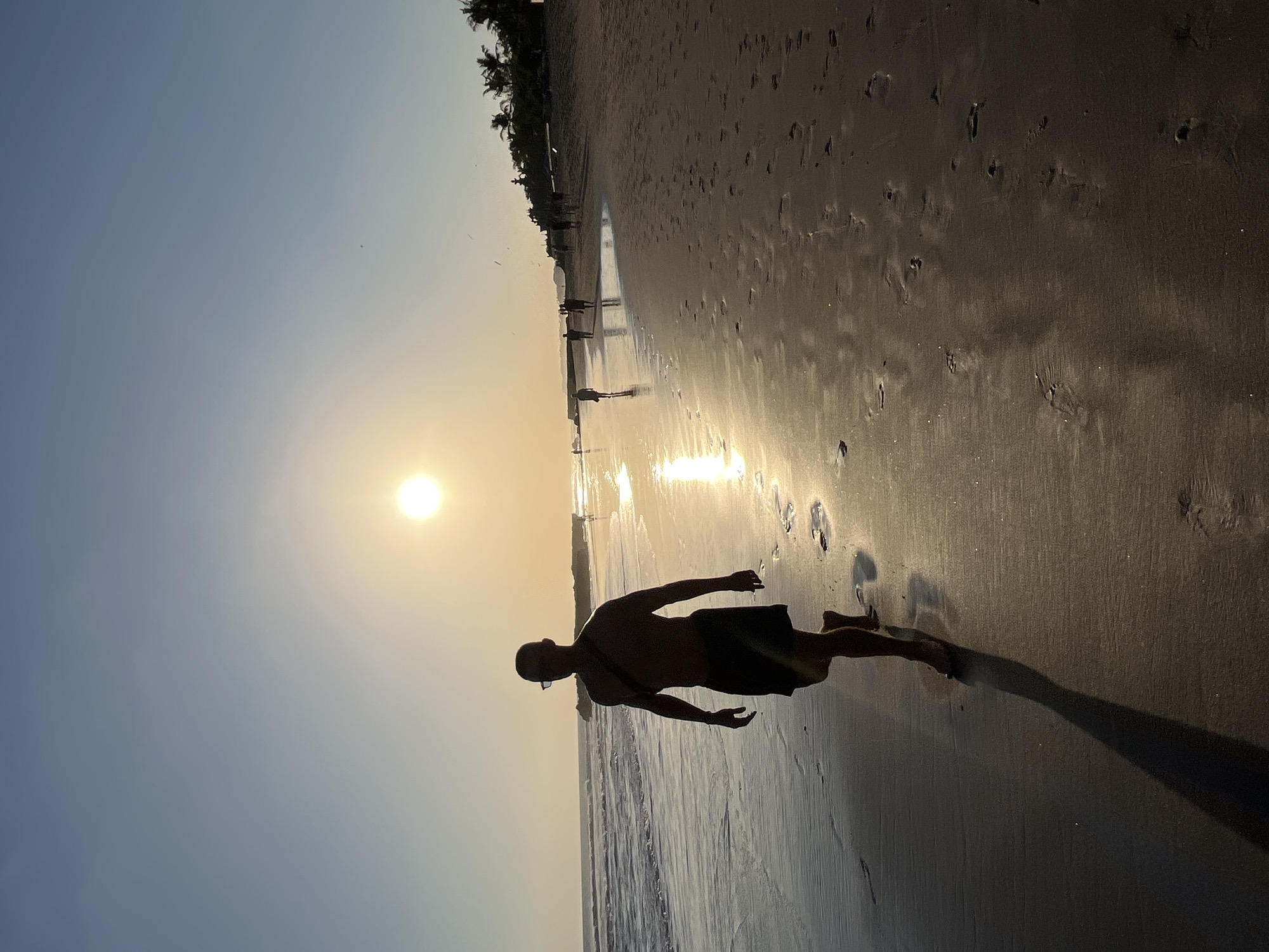 A man walks along a sandy beach in North Kuta, Indonesia. The sun is low in the sky, casting a golden glow and long shadows. The man is silhouetted against the bright sun, wearing dark shorts and a strap across his chest. Waves lap at the shore to his left, and footprints mark the wet sand. Other people can be seen in the distance walking along the beach. Palm trees line the edge of the beach in the background, with a hint of buildings or structures further inland. The scene conveys a sense of tranquility and the end of a day.