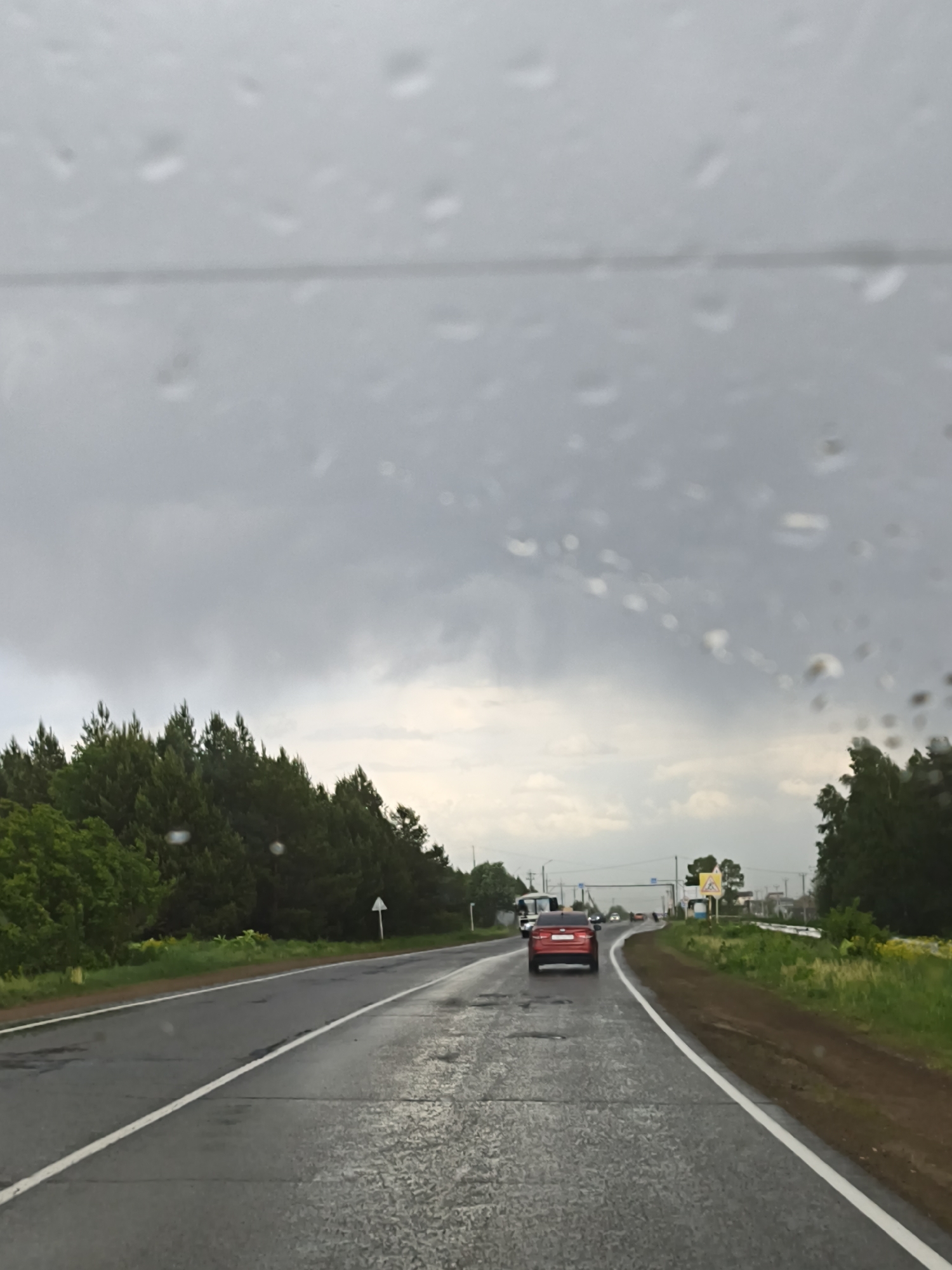 The image, taken from inside a vehicle through a rain-streaked windshield, depicts a wet road in the city of Прикамский, Russia. A red car is visible ahead on the road, along with other vehicles in the distance. The road is bordered by trees and vegetation on either side. The sky is overcast and cloudy, suggesting a rainy or stormy weather condition. A road sign is present along the roadside.