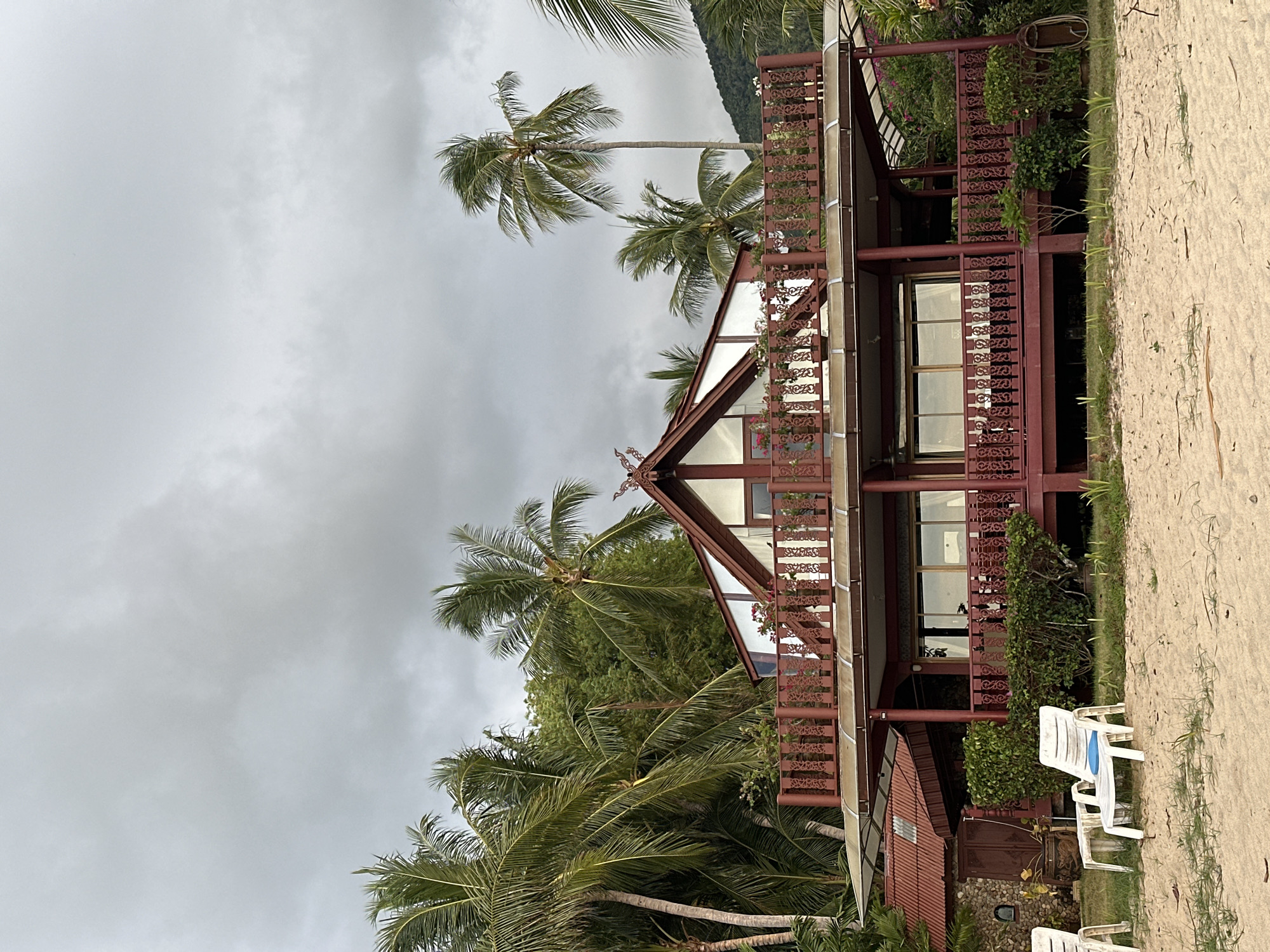 The image depicts a two-story, dark red wooden building with a steeply pitched roof and a large balcony on the upper level, situated on a sandy beach. The building is surrounded by lush green palm trees and dense foliage. The sky is overcast with gray clouds. In the foreground, two white plastic lounge chairs and a small table are placed on the sand, suggesting a relaxed, tropical setting. The architecture of the building, with its ornate railings and roof trim, hints at a Southeast Asian influence, consistent with the location of Baan Tai, Thailand. The overall scene conveys a sense of a beach resort or a private residence in a tropical climate.
