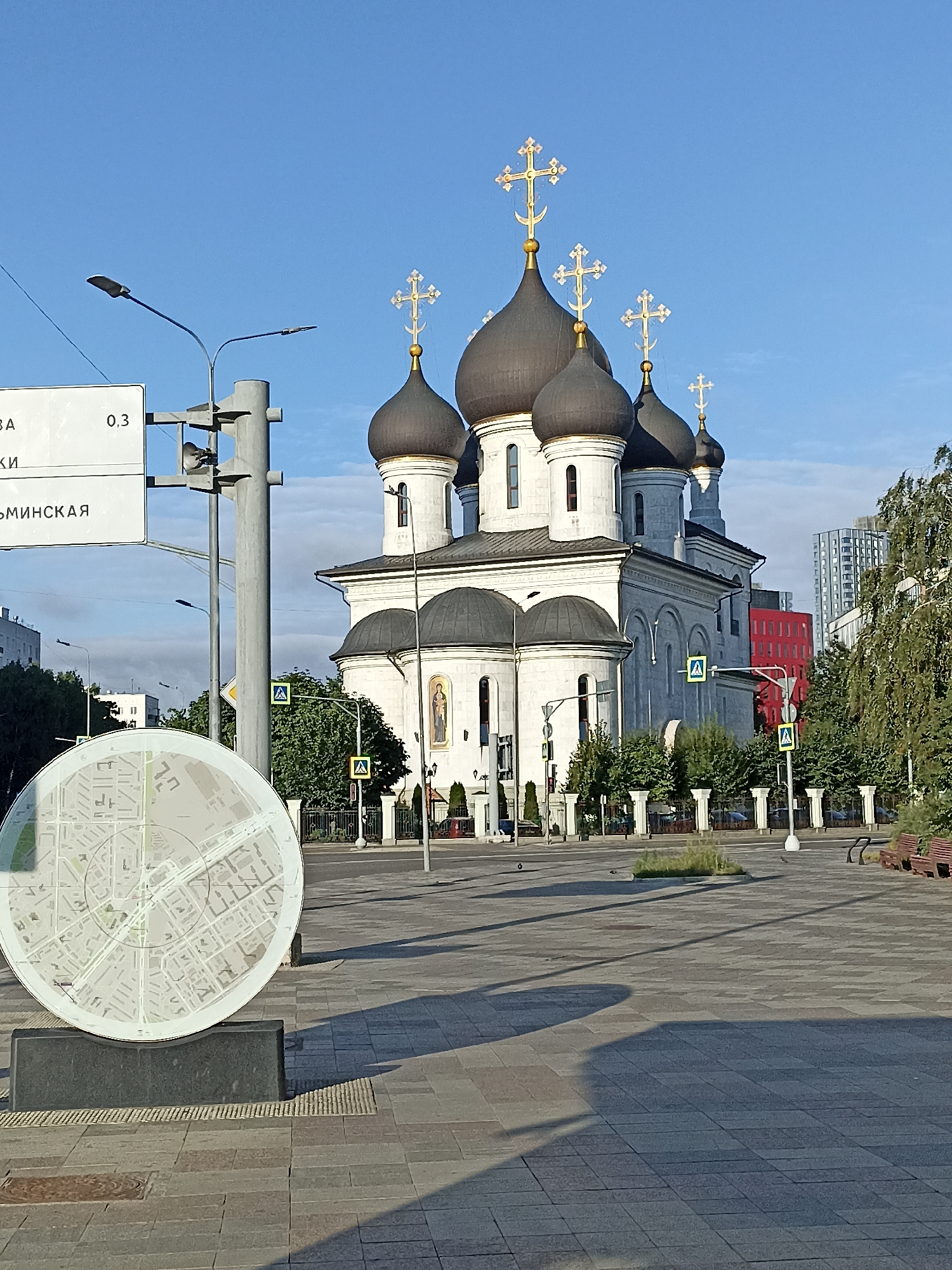 This is an exterior, eye-level, daylight photograph of the Church of the Resurrection of Christ in Moscow, Russia. The church is a large, white, Orthodox Christian building with multiple domes, topped with gilded crosses. It is situated in a paved plaza, with trees and modern buildings visible in the background. A circular information kiosk with a map of the area is positioned in the foreground to the left. Traffic lights and street signs are also visible, indicating this is an urban environment. The sky is clear and blue, suggesting fair weather. No people are visible in the photograph.