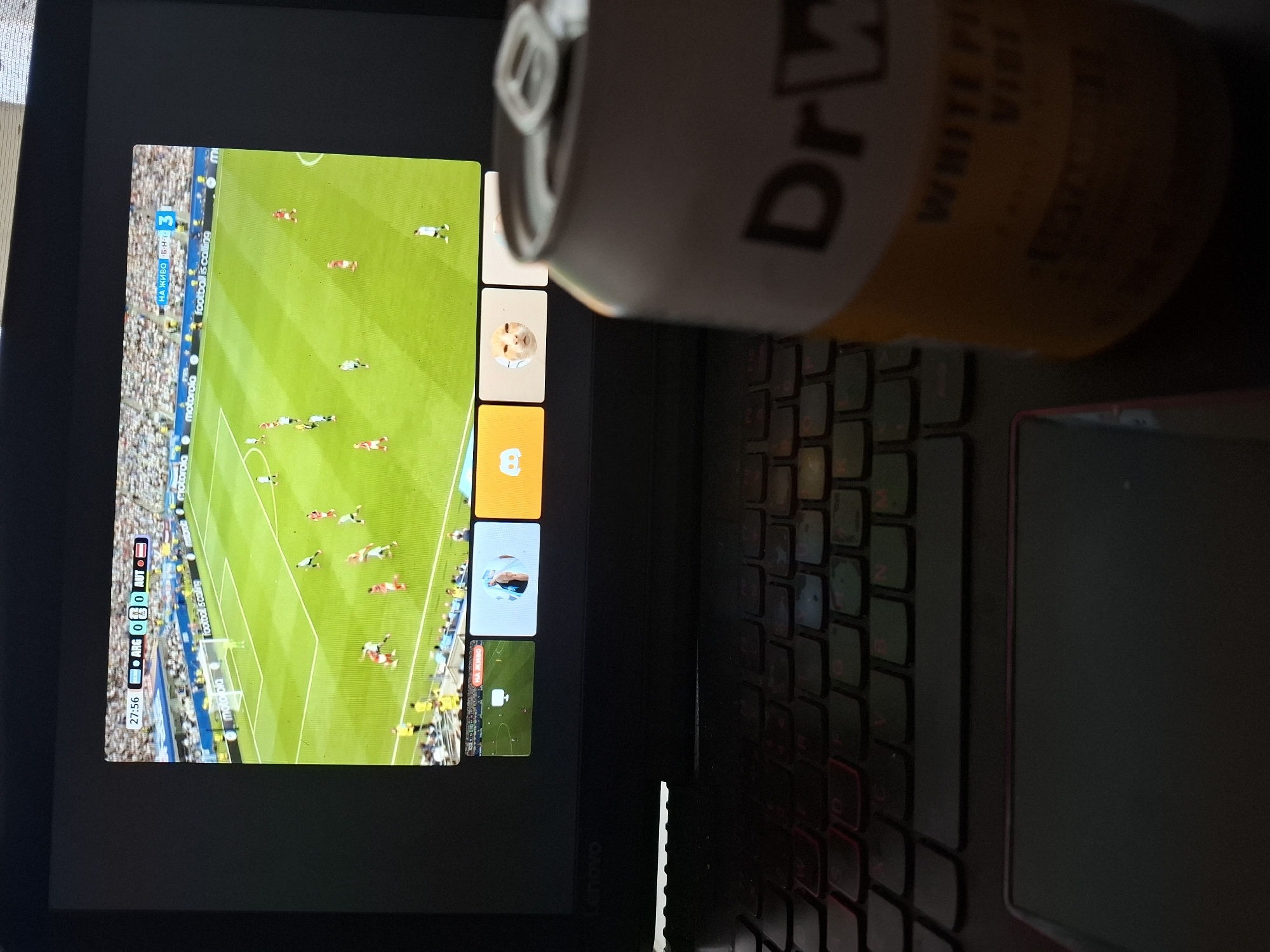 This image shows a close-up view of a laptop screen displaying a live football match. The screen is on the left side of the image, and a can of "Dr. W." drink is positioned in the foreground, obscuring part of the screen and keyboard. The football match appears to be between two teams, with the score showing "ARG 0 AUT 0". The time displayed on the screen is "27:56".
On the left side of the screen, there's a score graphic indicating "НА ЖИВО БНТ", suggesting the broadcast is live on BNT, a Bulgarian national television channel. The field is green, with players in red and white kits visible. There are also smaller preview windows or control elements on the right side of the main match display, showing icons and player images.
The background shows a portion of a laptop keyboard, with some keys illuminated with red backlighting. The overall setting appears to be indoors, likely in a room with subdued lighting. The presence of the drink can suggests a casual viewing experience. The orientation of the image is rotated, with the screen appearing to be viewed from a high angle. The text "motorola" is visible on some elements of the screen's interface, likely indicating sponsorship or branding. The overall impression is a snapshot of someone watching a football game on their laptop, with a drink readily at hand.