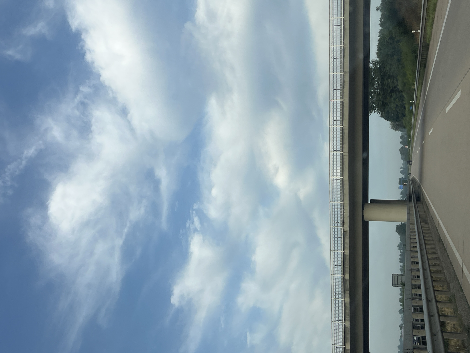 A view from a vehicle traveling on a highway in Unknown, Unknown. The sky is partly cloudy with scattered white clouds against a blue sky. A pedestrian overpass spans the highway in the mid-ground. The overpass is supported by a large cylindrical concrete pillar. The highway is a multi-lane road with a concrete barrier on the left side. Trees and vegetation line the sides of the highway in the background. The scene appears to be during the daytime.
