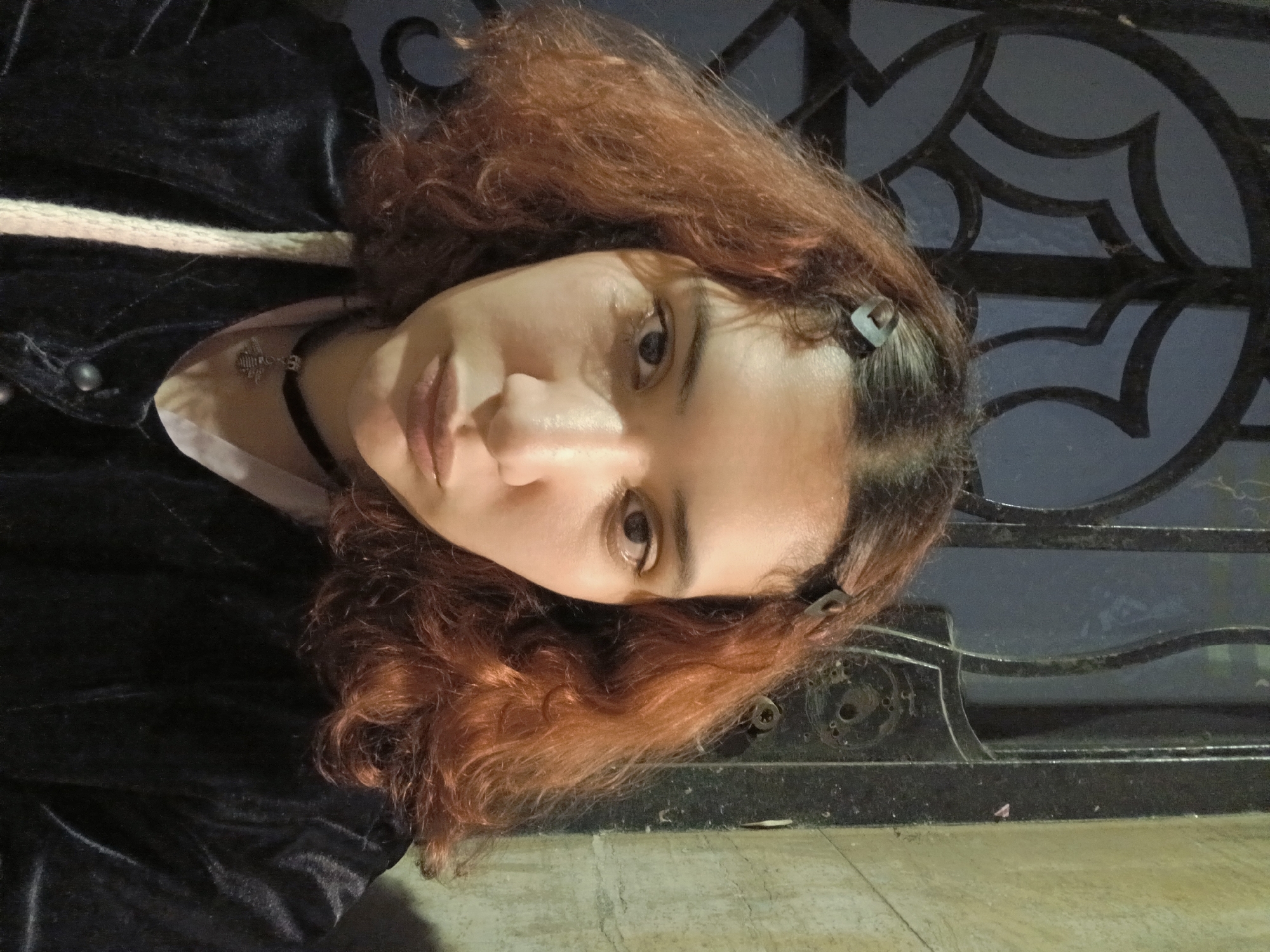 A young woman with reddish-brown, wavy hair is depicted in a head-and-shoulders portrait, her head tilted to one side as she gazes directly at the camera. Her expression is neutral and calm. Her brown eyes are prominent, and small dark hair clips are visible in her hair, pulling back some strands from her face.
She is dressed in a dark, possibly velvet, garment, with a lighter-colored collar or shirt visible beneath. A black choker adorned with a silver cross pendant is worn around her neck.
The background suggests an outdoor evening or nighttime setting in low light. Behind the woman is a dark, ornate metal fence or gate, featuring intricate, almost gothic, geometric patterns. This architectural detail could be a wrought-iron element common in cities like Buenos Aires, Argentina. Below the fence, a lighter-toned ground surface, possibly concrete or pavement, is visible. The ambient lighting casts soft shadows, highlighting the contours of her face. No visible text is present in the image.