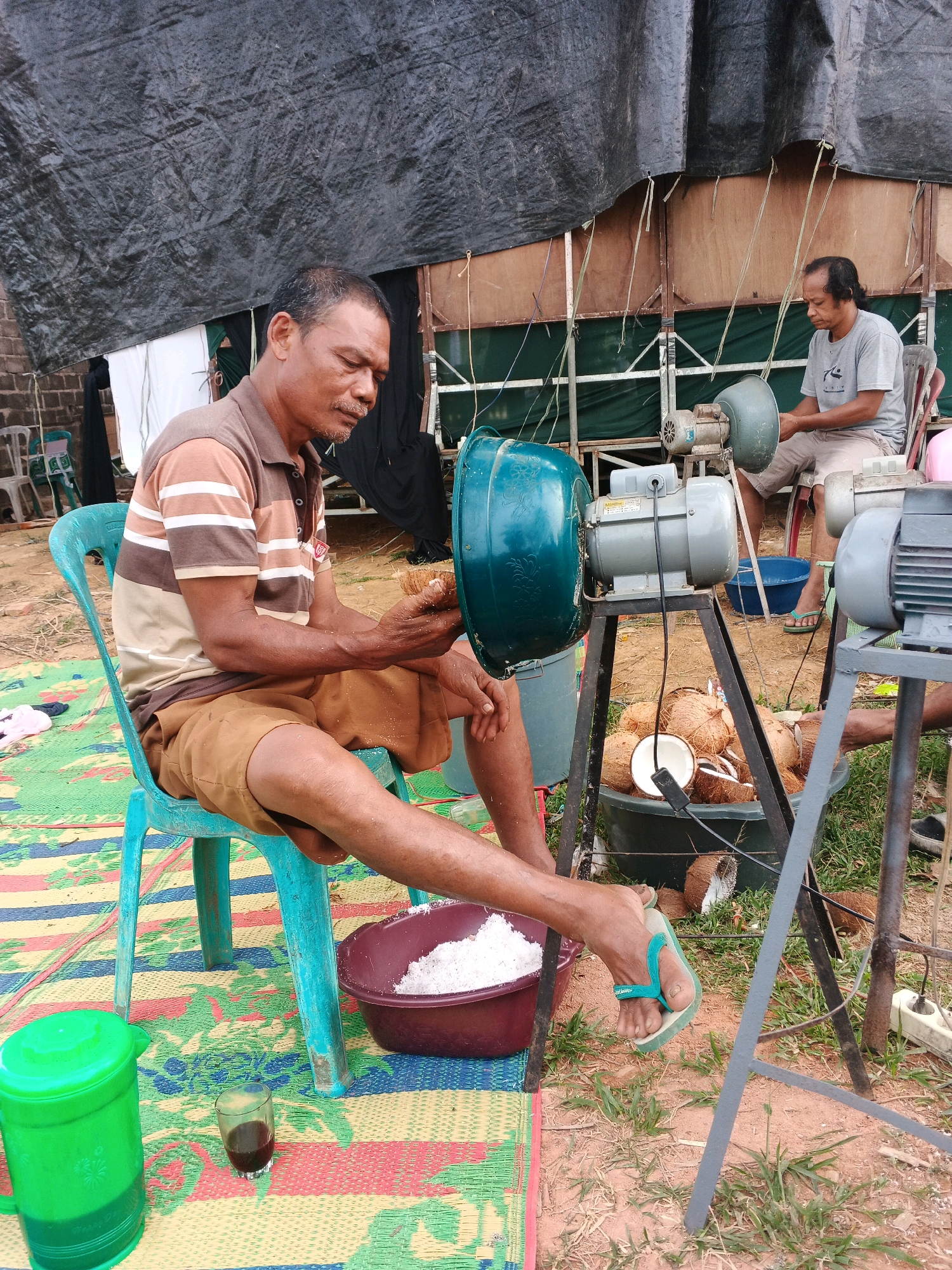 The image depicts an outdoor scene in Brangan Mulya, Indonesia, where two men are engaged in processing coconuts. In the foreground, a man sits on a turquoise plastic chair on a colorful woven mat. He wears a striped polo shirt and brown shorts. He is holding a coconut husk in his hands, appearing to prepare it for a machine. Next to him, on the mat, is a maroon bowl filled with shredded coconut and a small glass of dark liquid. To his left, a large green plastic container is visible.
Behind the man, an industrial-style coconut grater with a teal-colored casing and a motor is mounted on a metal stand. Further in the background, another man sits on a chair, also observing the process or perhaps waiting for his turn. He wears a grey t-shirt and light-colored shorts. Several coconuts, both whole and halved, are piled in a large dark tub in front of the grater. The area appears to be a makeshift workspace, possibly in a rural setting, with a black tarp overhead providing shade. The ground is covered in dirt and sparse grass. The overall impression is of a communal or small-scale commercial activity related to coconut processing.