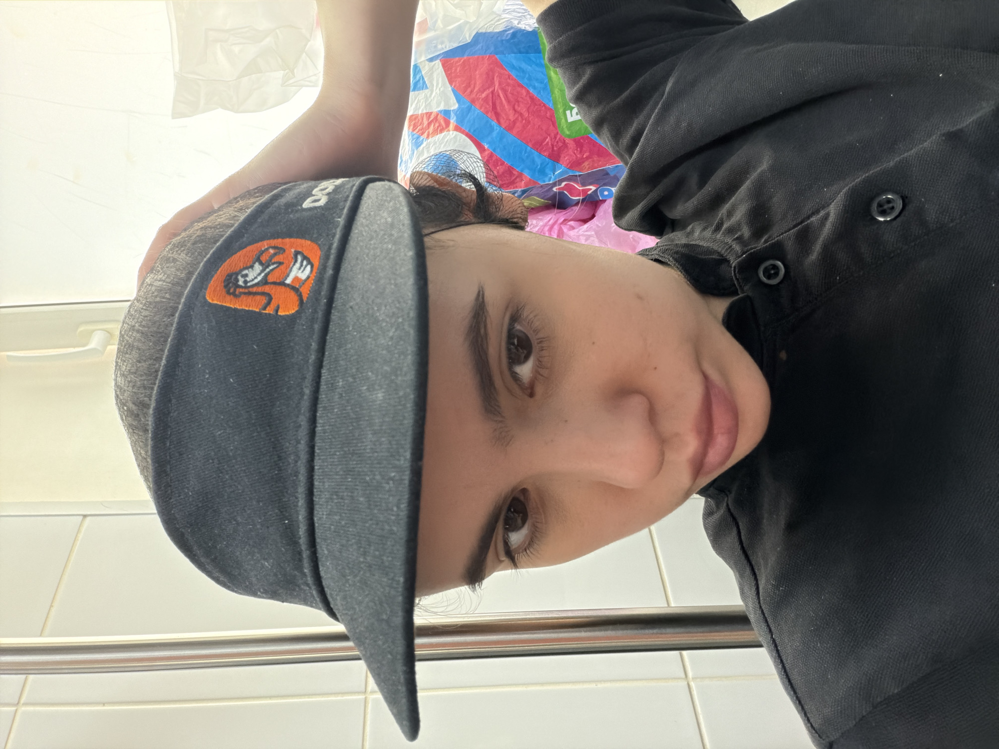 A close-up, canted, selfie-style shot depicts a person wearing a dark grey visor with an orange cartoon mascot on it and a dark-colored button-up shirt. The person is fair-skinned with dark eyebrows and brown eyes, looking directly at the camera. The background is out of focus but includes a shopping bag with colorful designs and what appears to be a white tiled wall with a metal handrail. There is no indication of Chaikovsky, Russia in the image.