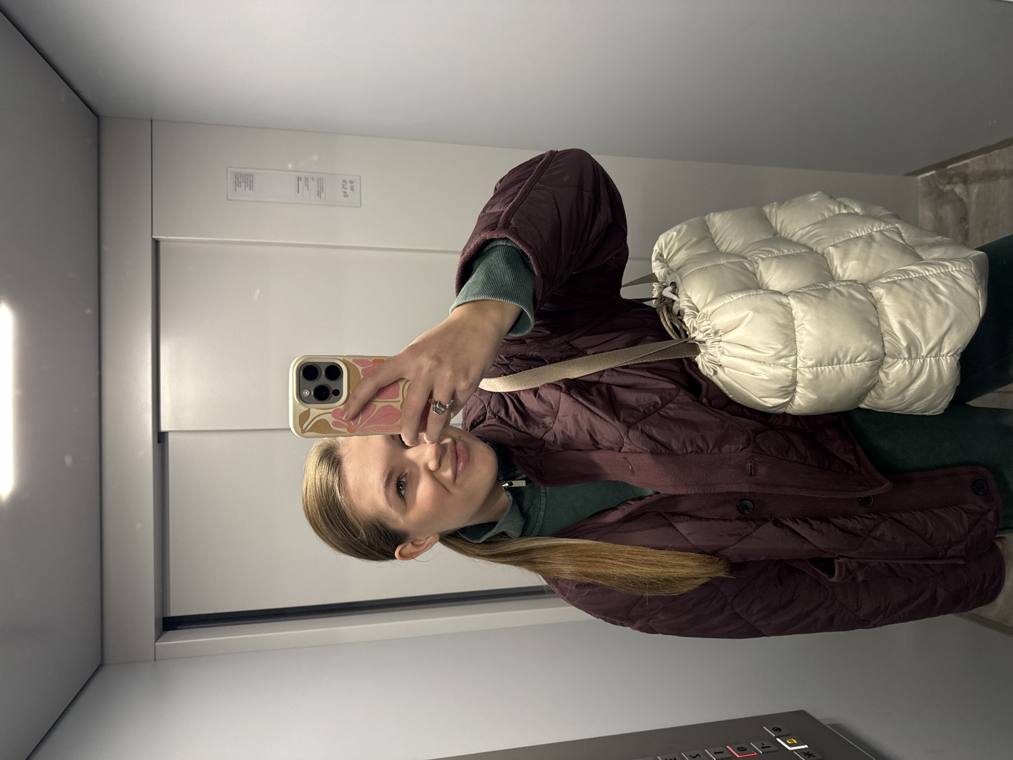 A young woman takes a selfie in an elevator. She is wearing a burgundy quilted jacket over a dark green top, and a cream-colored puffy drawstring bag hangs on her shoulder. Her hair is blonde and pulled into a low ponytail. The elevator interior is light grey and contains a control panel. The specific location in Vozdovac Urban Municipality, Serbia cannot be determined from the image.