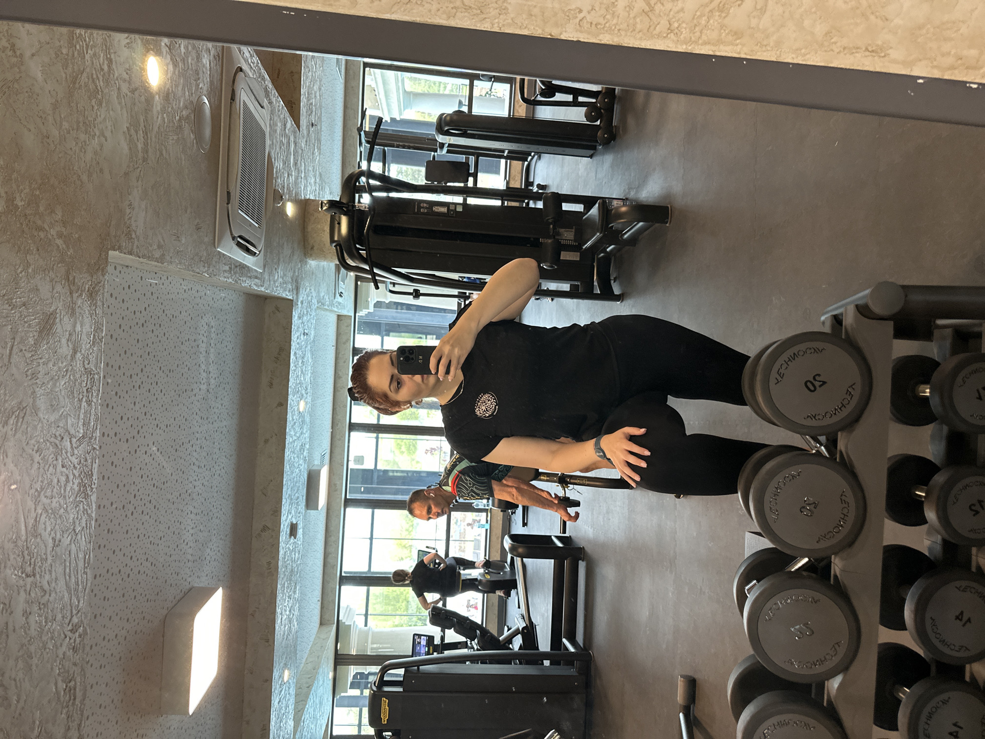 Here is a detailed description of the image:
The image shows the reflection of a gym interior in a mirror. A woman in black athletic wear is taking a selfie with her phone. In the background, other people are exercising on gym equipment. There are weight racks with dumbbells of various sizes visible in the foreground. The gym has large windows offering a view of the outside. The location appears to be in an urban area. There is no specific information in the image confirming that it is in Kazanlak, Bulgaria.
