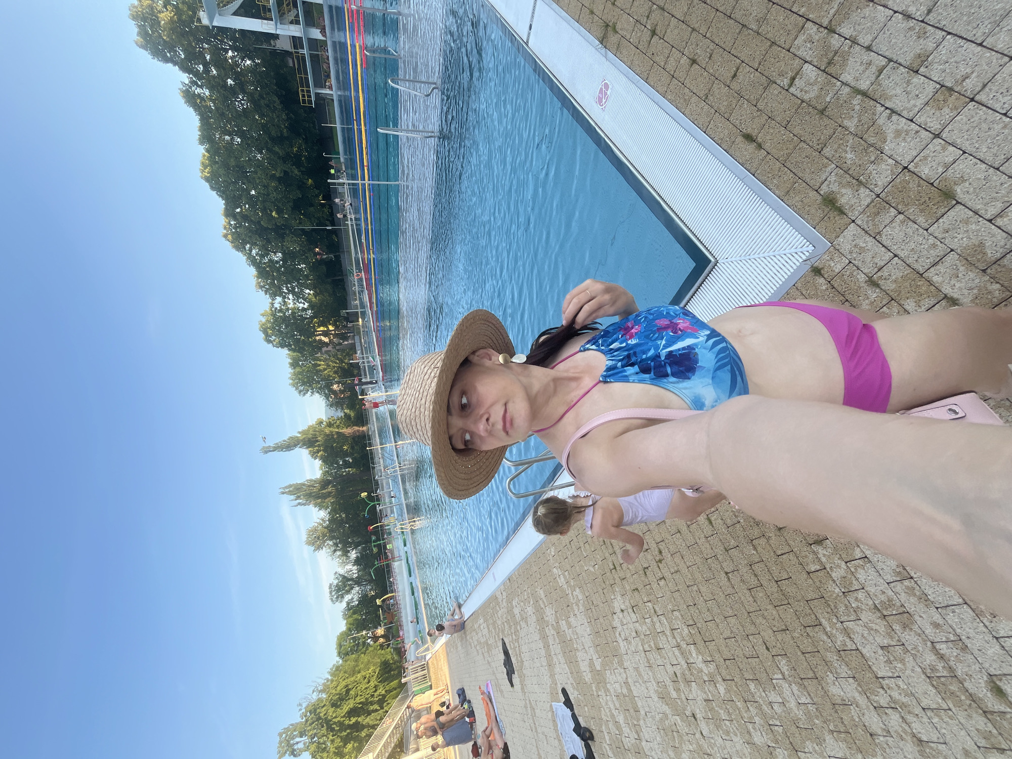 This media file shows a group of people at an outdoor swimming pool on a sunny day. The scene is dominated by a large blue swimming pool with lane markers. In the foreground, a woman wearing a straw hat and a blue and pink floral swimsuit is taking a selfie. She has a light pink strap from her swimsuit visible. Next to her, a young child in a light purple swimsuit is partially visible, facing away from the camera. The pool area has a brick-like paved surface, with a few towels and bags scattered around. In the background, there are trees and some structures associated with a swimming facility, possibly a diving board. The sky is clear and blue, suggesting good weather. The overall mood appears relaxed and recreational, indicative of a summer day at a pool. There are no discernible text elements in the image.