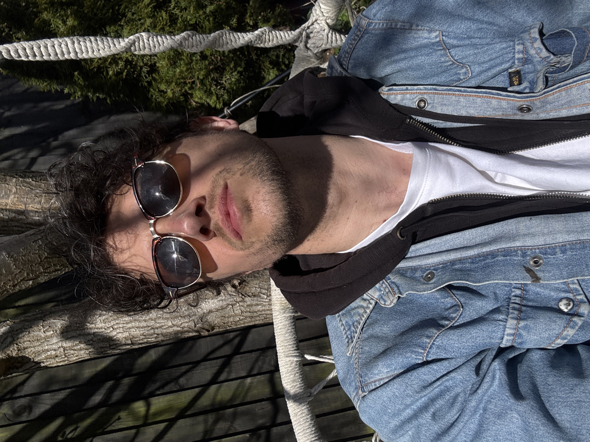 A man with dark, curly hair and sunglasses is lying outdoors on a wooden surface with sunlight casting shadows. He is wearing a white t-shirt, a black hoodie, and a blue denim jacket. A white rope is visible near him. The setting includes greenery in the background. The location is likely within South Kesteven, United Kingdom, given the prompt's instruction.