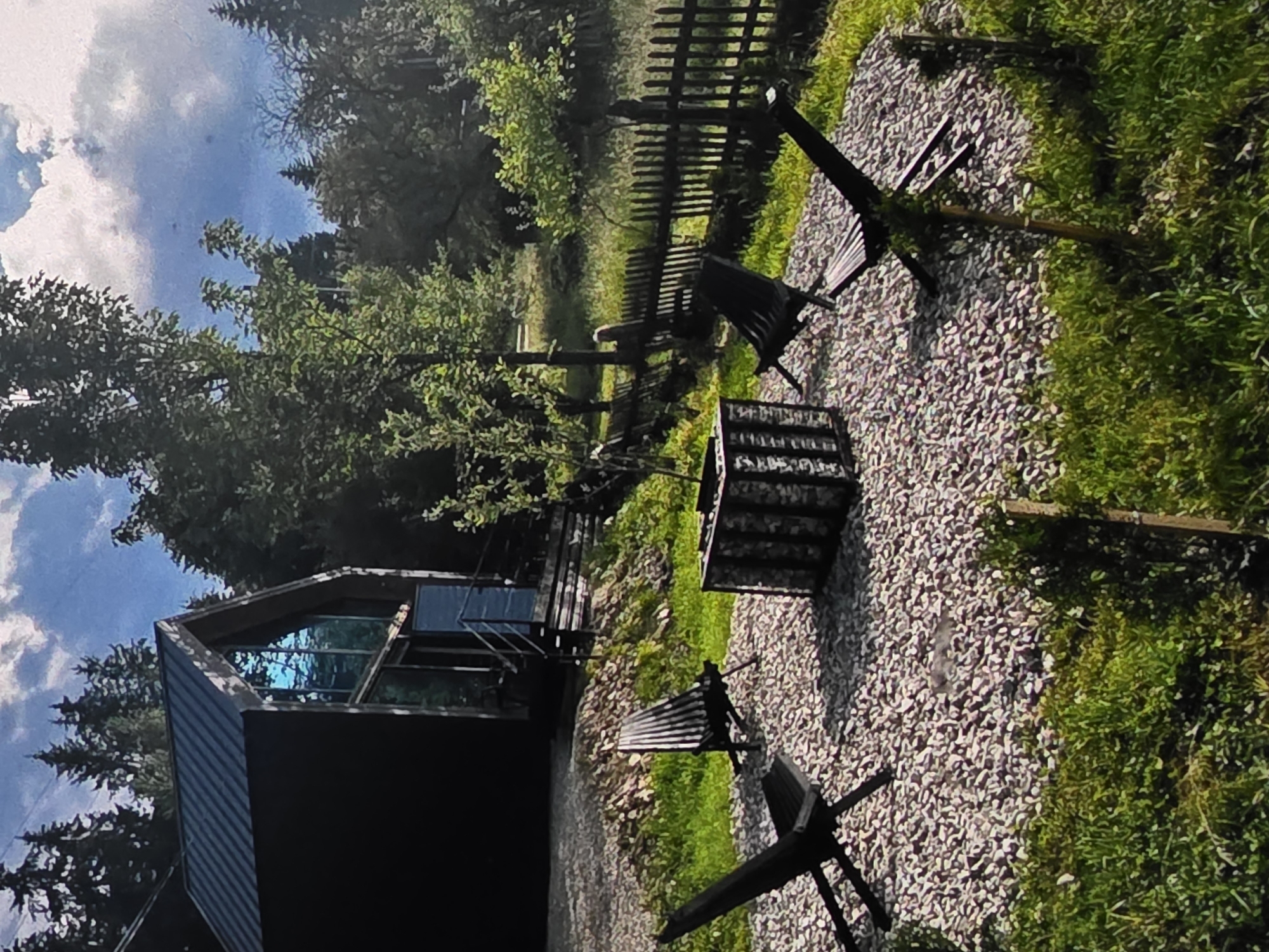 The image shows a modern black cabin with large glass windows, nestled amongst trees. In front of the cabin, there is a gravel area with a fire pit and two black chairs. A wooden fence runs along the edge of the property, separating it from a grassy area. The sky is blue with white clouds, suggesting it is daytime and the weather is pleasant. The scene appears to be a tranquil, secluded retreat, possibly in a rural or mountainous area, as indicated by the presence of dense trees and a rustic fence. The overall mood is one of peaceful relaxation and outdoor enjoyment. There is no visible text in the image. The provided location context is Dubno, Ukraine.