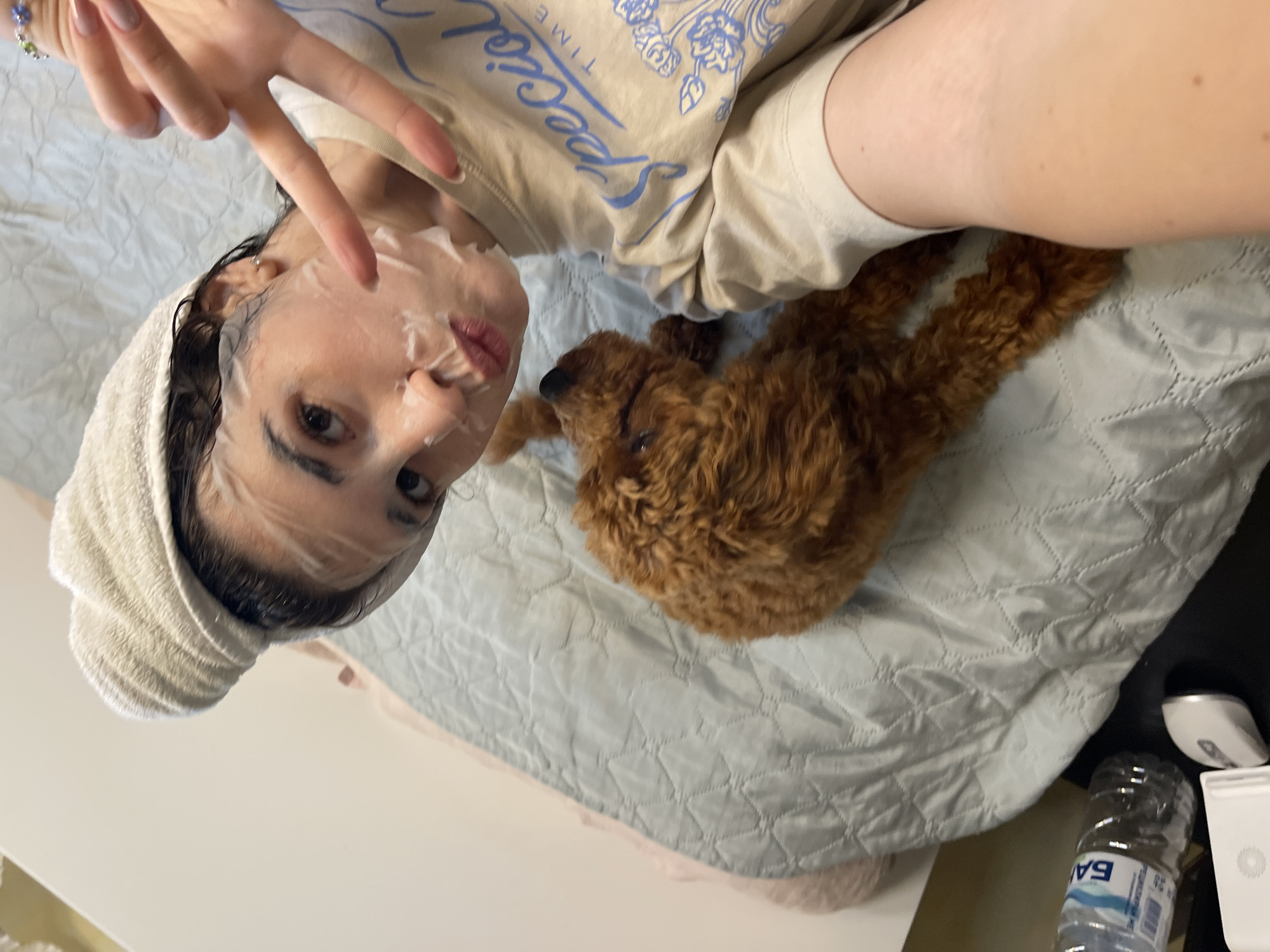 A young woman with a towel wrapped around her head and a sheet mask on her face is lying on a bed. She is making a peace sign with her right hand and pouting her lips. A small, brown poodle is lying next to her on the bed. A t-shirt with the words "Special Time" printed on it is visible on the woman. A water bottle, a white rectangular object, and a white circular object are on the surface next to the bed. The scene is indoors, likely in a bedroom.