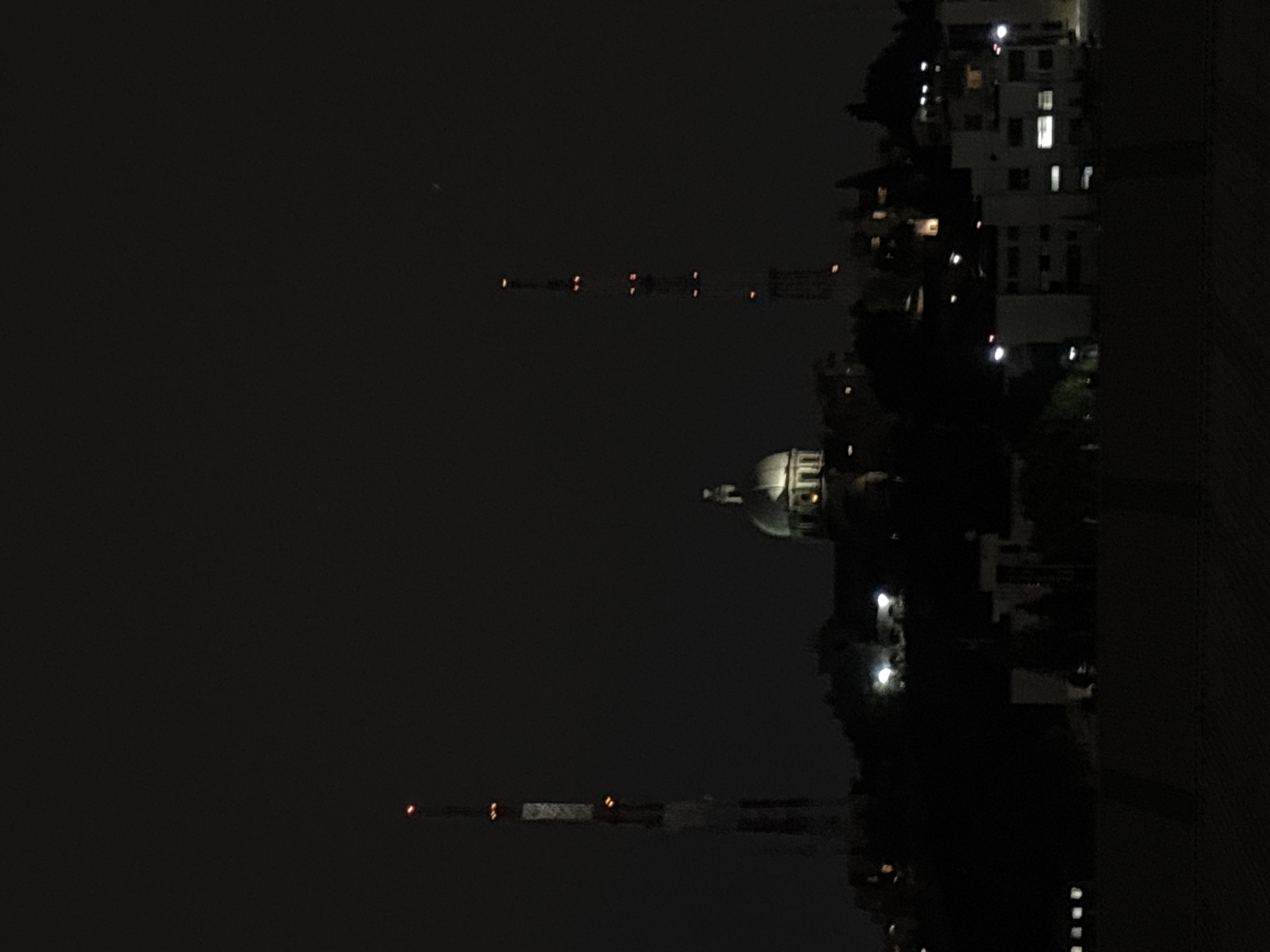 At night, the city of Puebla, Mexico, is dimly lit. In the distance, a building with a dome is illuminated, while to its left, two tall towers display blinking red lights. Further to the right, a multi-story building has a few lit windows. A dark vertical structure stands on the right. The sky is dark and unclear.