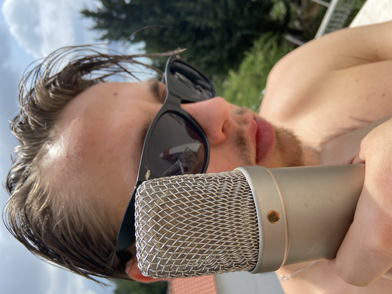 The image captures a close-up, low-angle perspective of a young person, likely male, reclining outdoors in Šušanj, Montenegro. The subject is shirtless and appears to have damp or wet, light brown hair styled back from their forehead. They are wearing dark, rectangular sunglasses that obscure their eyes.
The central focus is a large, professional-grade condenser microphone, held by the person's right hand. The microphone features a silver mesh grille and a light-colored, cylindrical body with a small gold accent screw visible on its side. The person holds the microphone close to their mouth, which is slightly open, revealing their lips. They have light facial hair, appearing as stubble or a short beard, around their chin and upper lip. A thin silver chain is visible around their neck.
The background shows a bright, clear blue sky with some scattered white clouds on the left side, transitioning to lush green foliage, likely trees, on the right. This suggests a sunny outdoor setting, possibly in a garden or by a poolside, under a Mediterranean climate. The strong sunlight indicates it is daytime, likely mid-day. The scene conveys a relaxed, casual atmosphere, possibly an outdoor recording session or vocal practice. There is no visible text in the image.