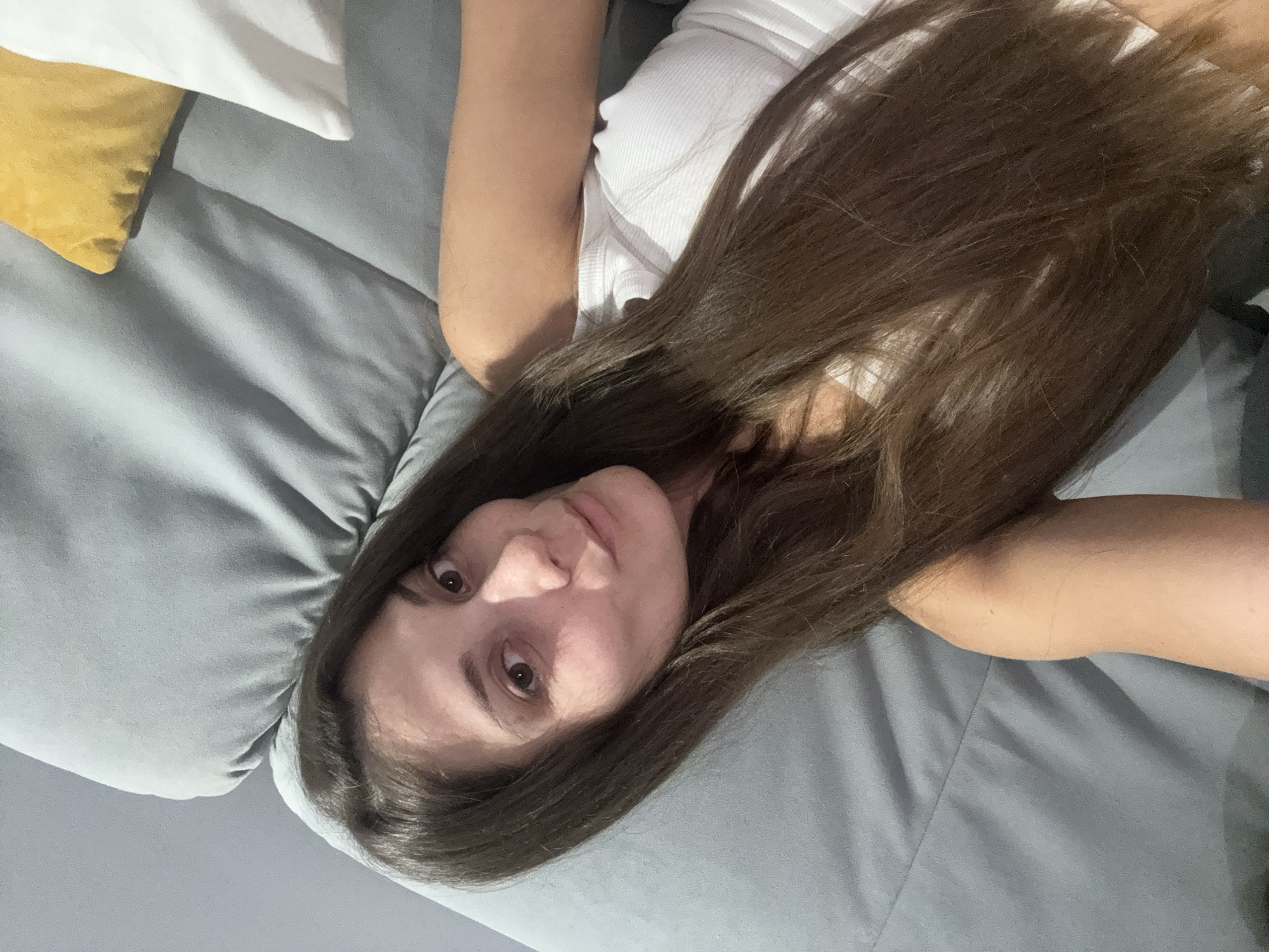 A young woman with long, brown hair is lying on a grey sofa, taking a selfie. She is wearing a white tank top. Her face is turned towards the camera, and she has a slight smile. The background of the image shows the grey sofa with a yellow and a white pillow visible on the right side. The lighting suggests an indoor setting. There is no discernible information about the location in Burgas, Bulgaria, in the image itself.