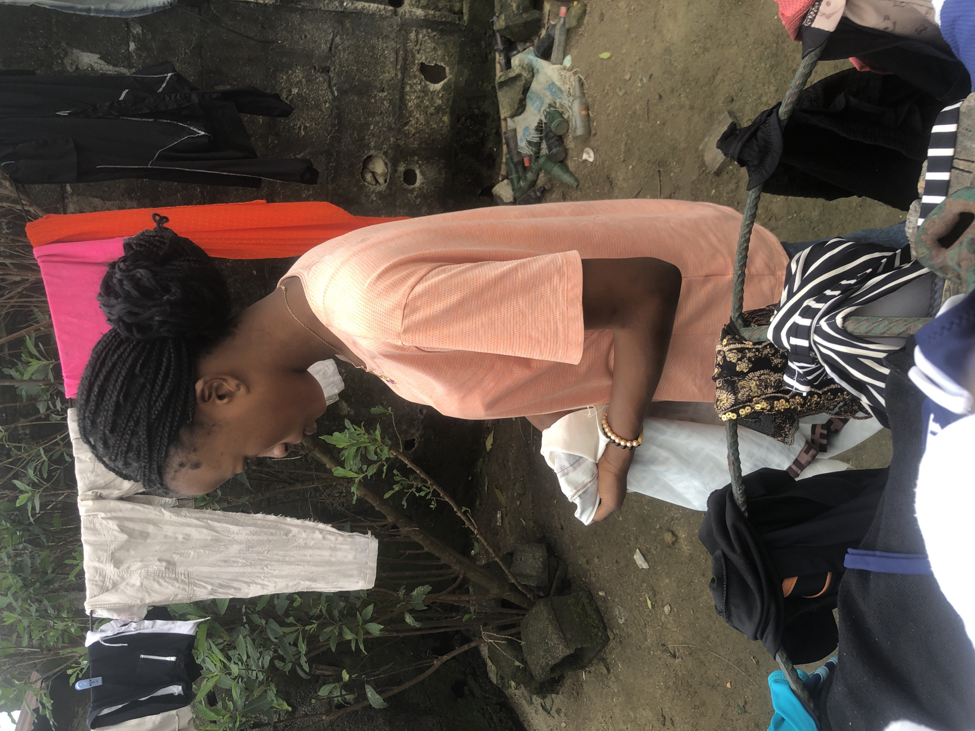 A young woman with braided hair pulled into a bun stands outdoors, tending to laundry. She is wearing a coral-colored t-shirt and jeans. She is holding a white garment in her hands. Various items of clothing are hung on a clothesline in the background, including a pink and an orange fabric, and black garments. To the left of the woman, a small tree with green leaves grows from behind a brick structure. The ground is dirt and scattered with debris, including plastic bottles and pieces of fabric. The scene appears to be in an outdoor laundry area, possibly in a residential courtyard in Ado, Nigeria.