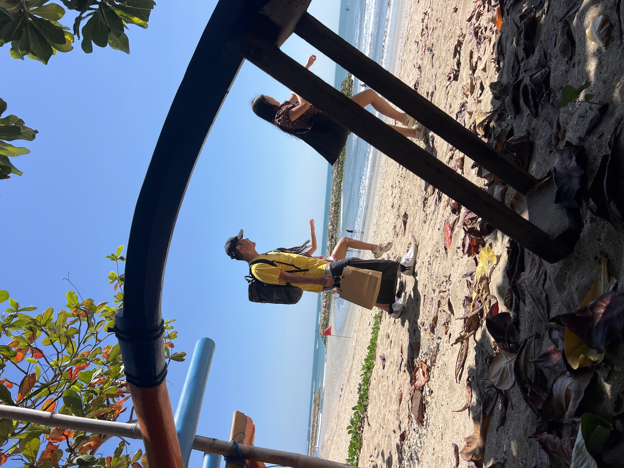 In Kuta, Indonesia, three individuals walk along a sandy beach under a clear blue sky. In the foreground, a dark, curved structure partially obstructs the view, with scattered dry leaves on the sand. Beyond this, a man in a yellow t-shirt and a black baseball cap, carrying a backpack and a brown paper bag, walks alongside a young girl in a patterned top. A second girl, wearing a dark polka-dot dress, walks slightly ahead of them. The background features the ocean with gentle waves, a stone breakwater, and a red flag planted in the sand. Overhead, green and reddish leaves of a tree are visible against the sky. The scene appears to be set on a sunny day.