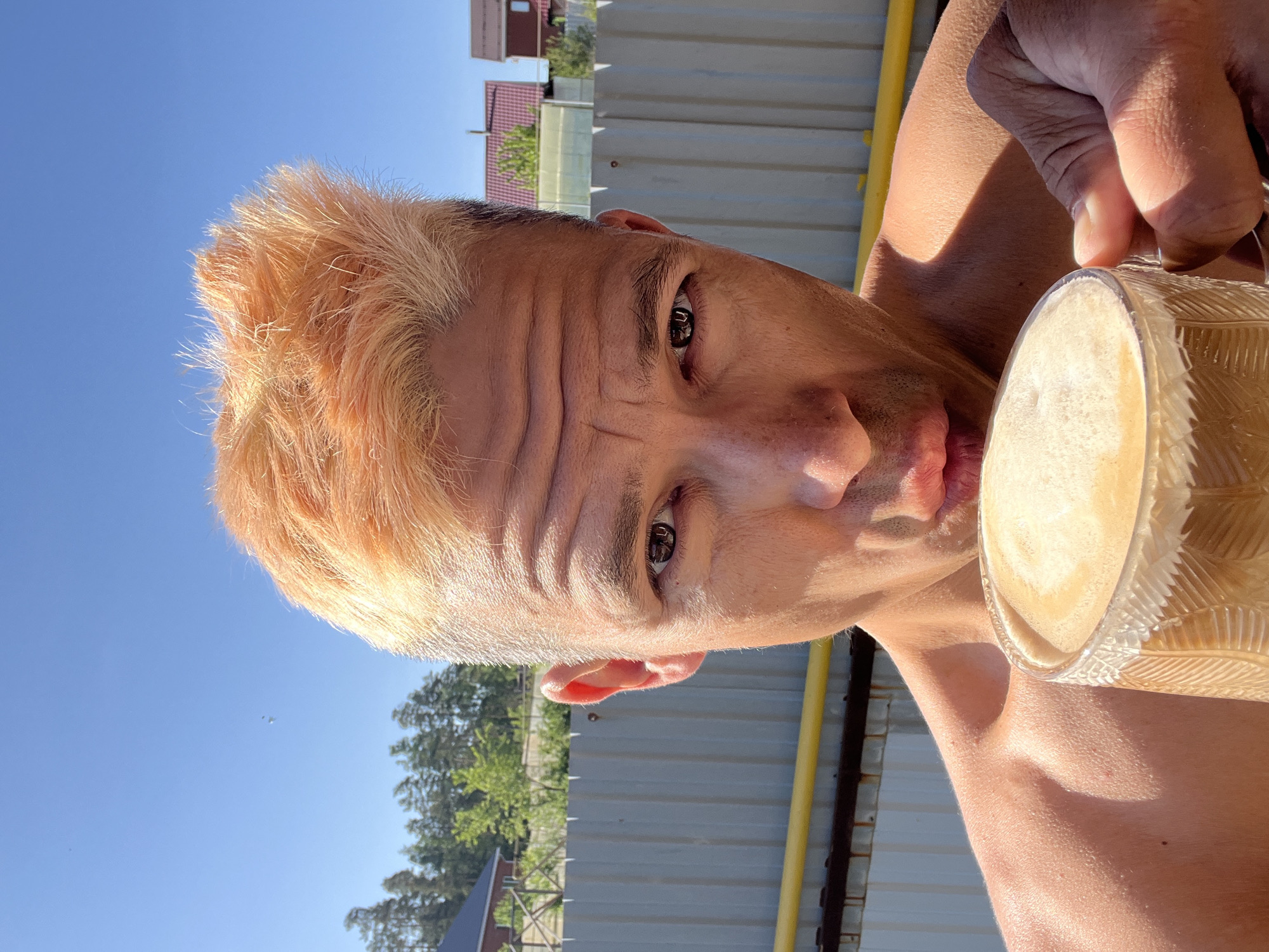 A man with dyed blonde hair is shown from a close, low-angle perspective, as he appears to be drinking from a glass. The setting is outdoors, with a blue sky partially visible. A fence, trees, and a building with a red roof are visible in the background, which may be located in Chaikovsky, Russia. The man's face is expressive, with visible forehead wrinkles. The glass appears to contain a light-colored liquid, possibly a beverage. The light suggests it is daytime.