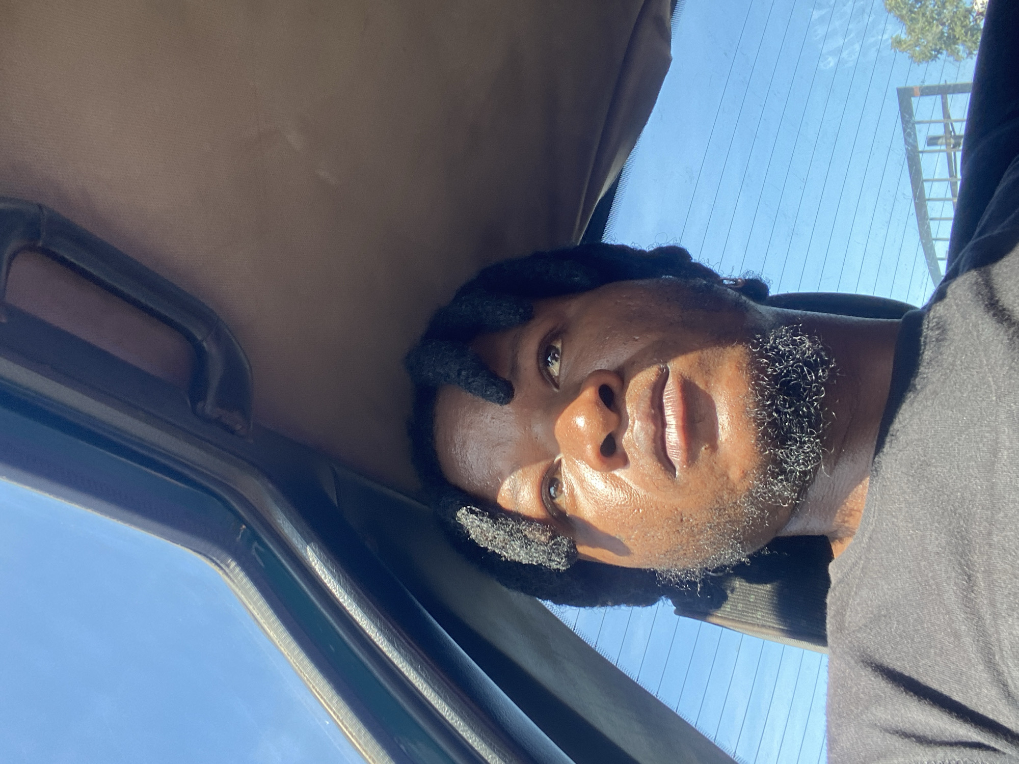 A medium close-up shot captures a Black male with dreadlocks and a goatee looking upwards, with sunlight illuminating his face. He is seated in what appears to be the interior of a vehicle, possibly a car or bus, with a sunroof open to a bright blue sky. The interior of the vehicle is visible, featuring a brown headliner and dark trim. The man is wearing a black t-shirt. The background seen through the car window shows a clear blue sky with faint horizontal lines, suggesting a car window with defroster elements. Further in the distance, a metallic structure and some greenery are visible, indicating an outdoor urban or suburban environment. There are no discernible landmarks in the image that specifically indicate the city of Jos, Nigeria.