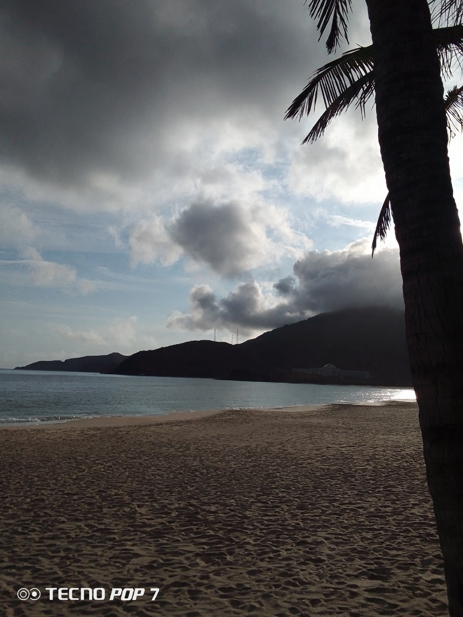 The image depicts a beach scene, likely near Pedro González, Venezuela, given the hints. A sandy beach stretches across the foreground, meeting a body of water. In the midground, there's a coastline with hills or low mountains silhouetted against the sky. A building is visible on the coast near the base of a hill. The sky is overcast with clouds, some of which are dark and heavy, while others are lighter. The sun appears to be shining through a break in the clouds. A palm tree trunk occupies the right side of the frame. The lower left corner contains the text "TECNO POP 7".