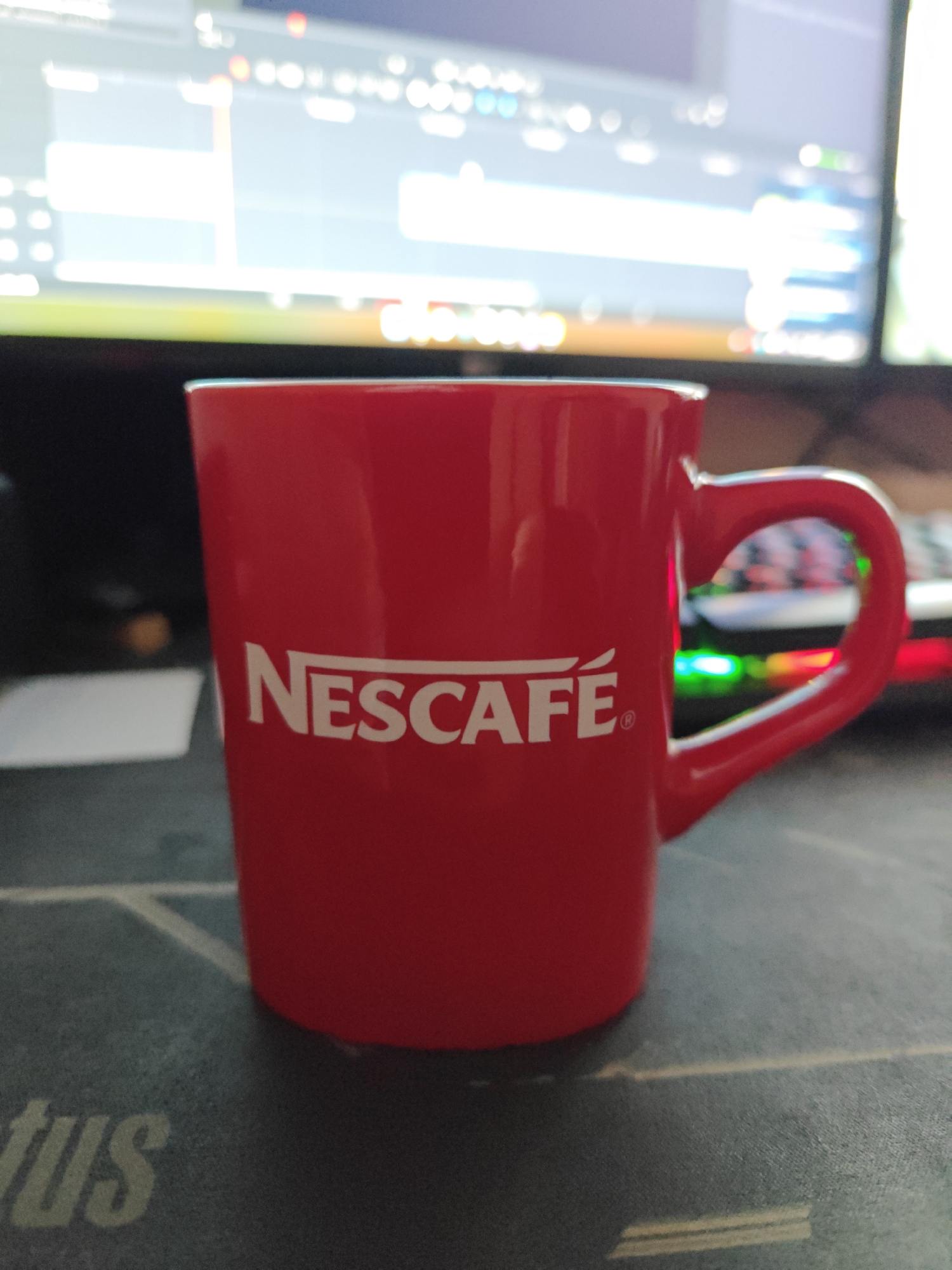 A red Nescafé mug is the primary subject, placed on a textured dark surface. The mug has a white logo that reads "NESCAFÉ". The background is blurred but appears to be a computer monitor displaying some kind of software interface, with colored lights from what might be a keyboard visible behind the mug. The image is taken from a slightly elevated perspective. The scene is indoors and does not provide any specific information about the city of Chaikovsky, Russia.