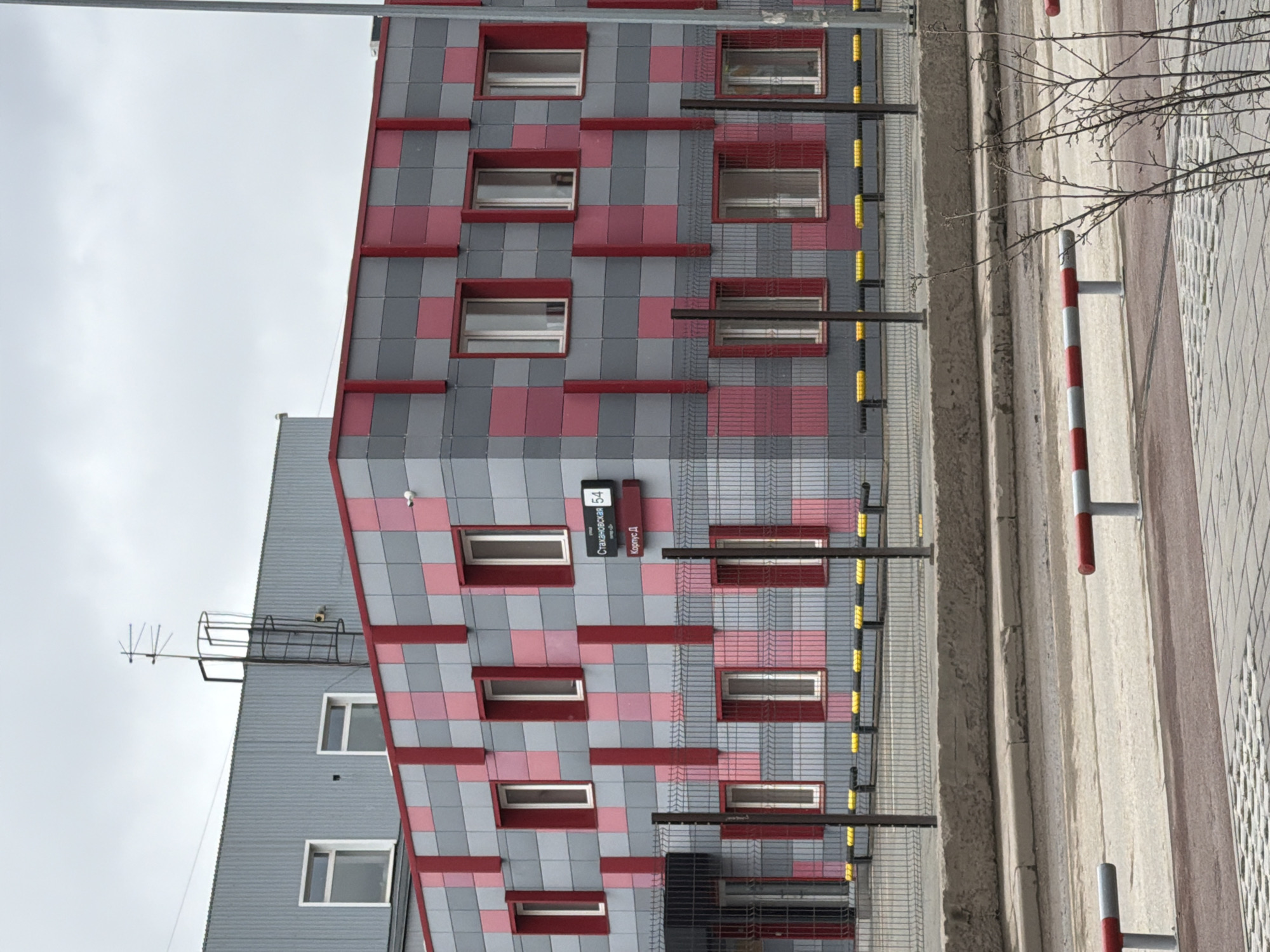 The image shows a building facade with a modern design, featuring a gray and red checkered pattern. The building is in Perm, Russia, based on the signage on the building. The building has a series of rectangular windows framed in red. Some of the windows are partially obscured by a metallic grid. A smaller section of the building has a gray facade and visible antennas. Along the bottom of the image, a sidewalk with red and white striped bollards is visible, and a leafless tree branch extends into the scene. The sky is overcast.