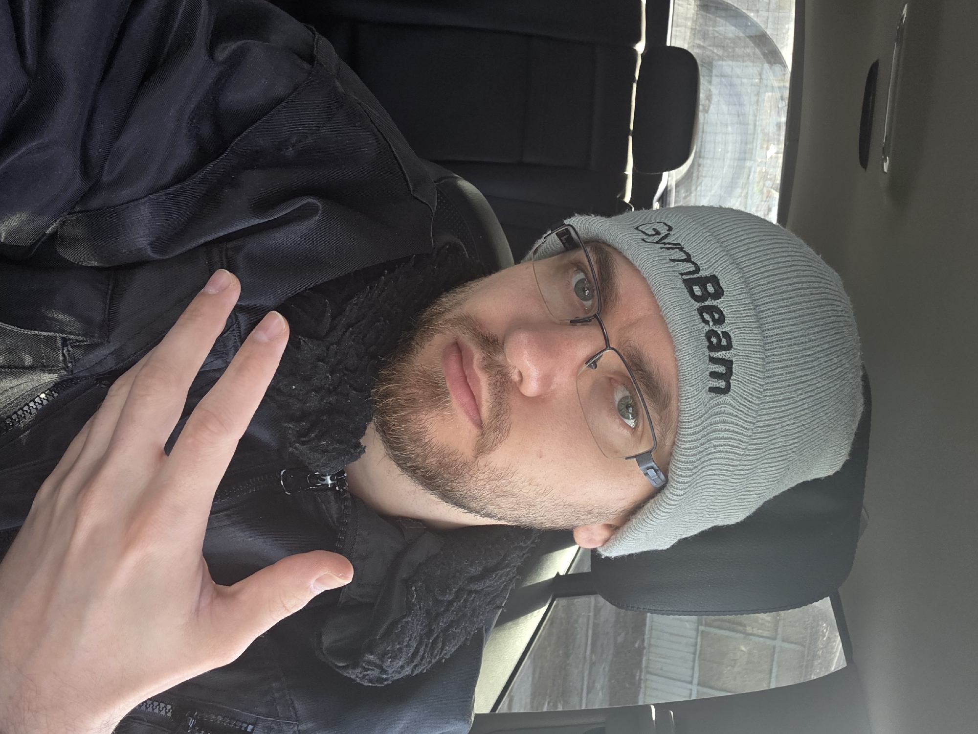 A man is photographed in the interior of a car. He is wearing a gray beanie with the word "GymBeam" printed on it, and glasses. He has a short beard. He is also wearing a black jacket. His left hand is visible in the foreground. The interior of the car is dark, with black seats. Outside, there appears to be a window showing a glimpse of an urban environment. The location is unidentifiable as Plovdiv, Bulgaria.