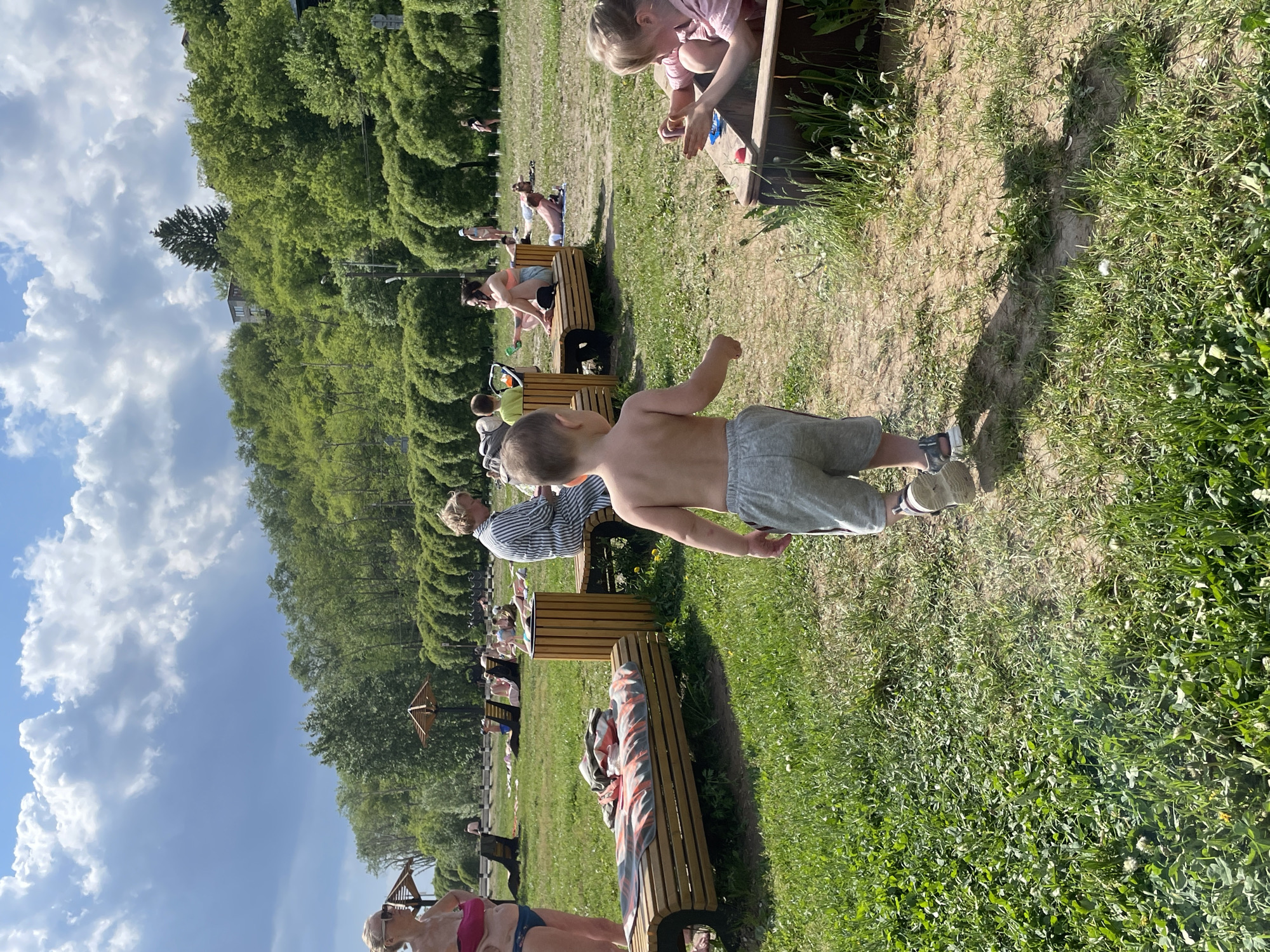 The image shows a sunny day at a park, possibly a city beach, with people relaxing and children playing. Several individuals are seated on wooden benches along a path. A young boy, shirtless and wearing shorts and sneakers, walks in the foreground. In the background, a row of trimmed trees lines the area. There are people sunbathing on the grass. The location is likely in Perm, Russia, based on the presence of characteristic vegetation and architectural styles.