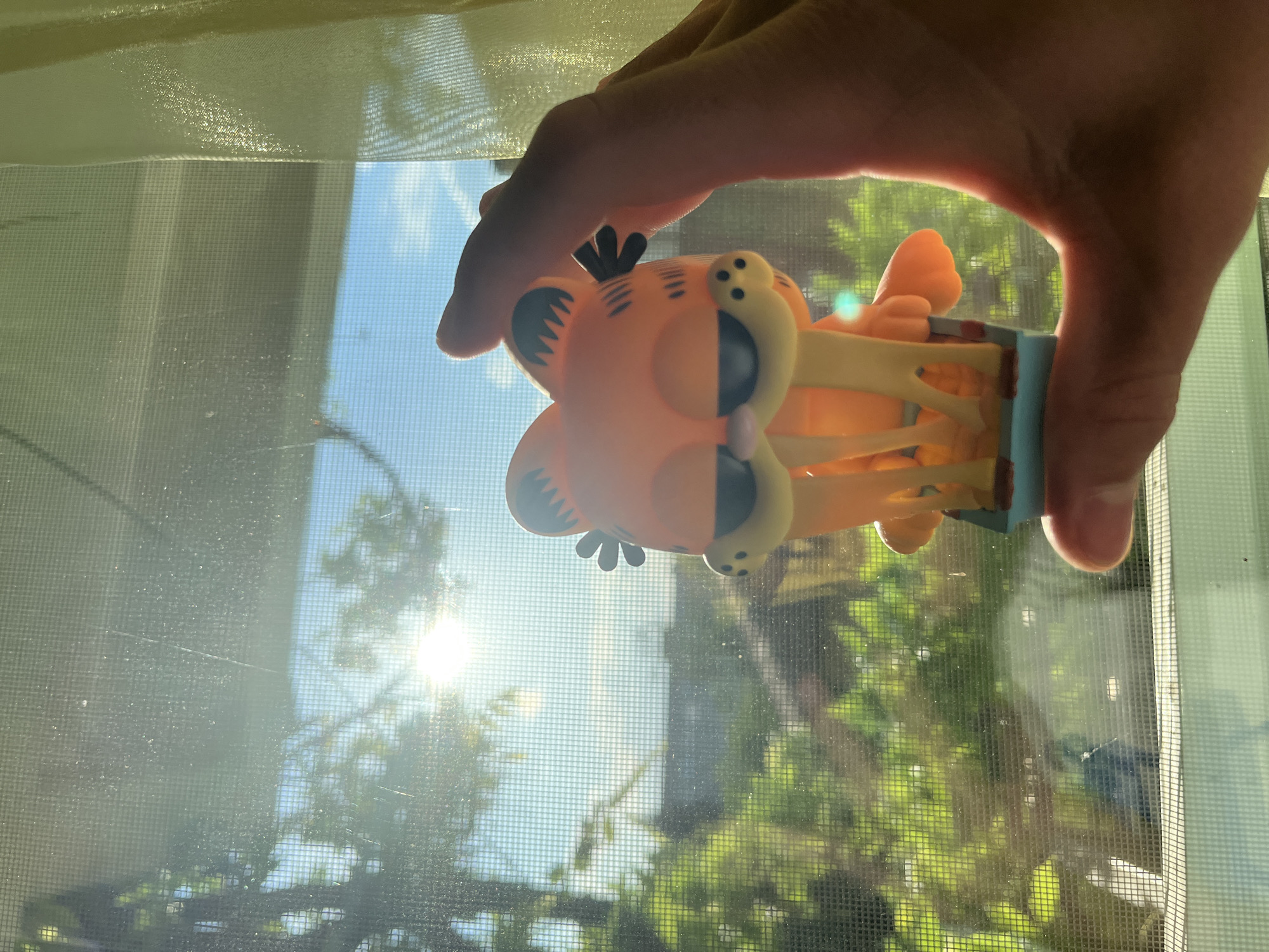 An eye-level, vertically oriented photograph captures a hand holding a Garfield figurine against a sunlit window in Sofia, Bulgaria. A light-skinned hand, illuminated from behind, occupies the top and right side of the frame, grasping an orange and yellow accordion-style toy. The toy distinctively depicts Garfield, the cartoon cat, with his recognizable head, black stripes, and ears. Its flexible body consists of ribbed, interconnected segments, and it rests on a blue and white striped base.
The scene is set against a window covered by a prominent mesh screen, visible across the entire frame with its grid pattern and small specks of dust. Through this screen, a bright outdoor environment is revealed. A clear blue sky is visible, particularly on the left, where the intense sunlight creates a strong lens flare and glare. Green foliage and trees are distinctly seen beyond the screen, indicating a natural landscape outside. The bright conditions suggest it is daytime, likely a clear and sunny morning or afternoon. The activity is simply the holding of the toy up to the window, possibly for play or to capture the light. There is no visible text in the image.