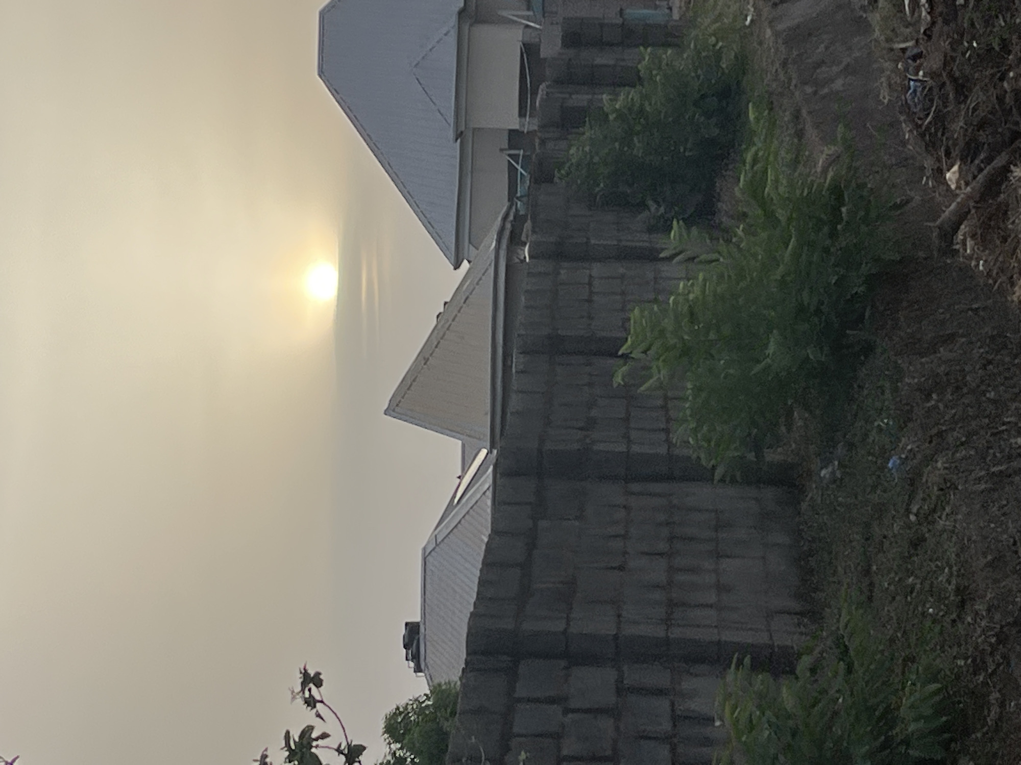 The image captures a scene in Abuja, Nigeria, during what appears to be either sunrise or sunset, given the low position of the sun in the sky and the warm, diffused light.
Foreground: The foreground is dominated by a partially constructed wall made of dark grey concrete blocks. Patches of green vegetation grow adjacent to the wall, and some weeds and debris are visible on the ground.
Middle Ground: Several structures, likely residential buildings, are visible behind the wall. They have white roofs and light-colored walls.
Background: The background consists primarily of the sky, which is hazy or dusty. The sun is visible as a bright orb.